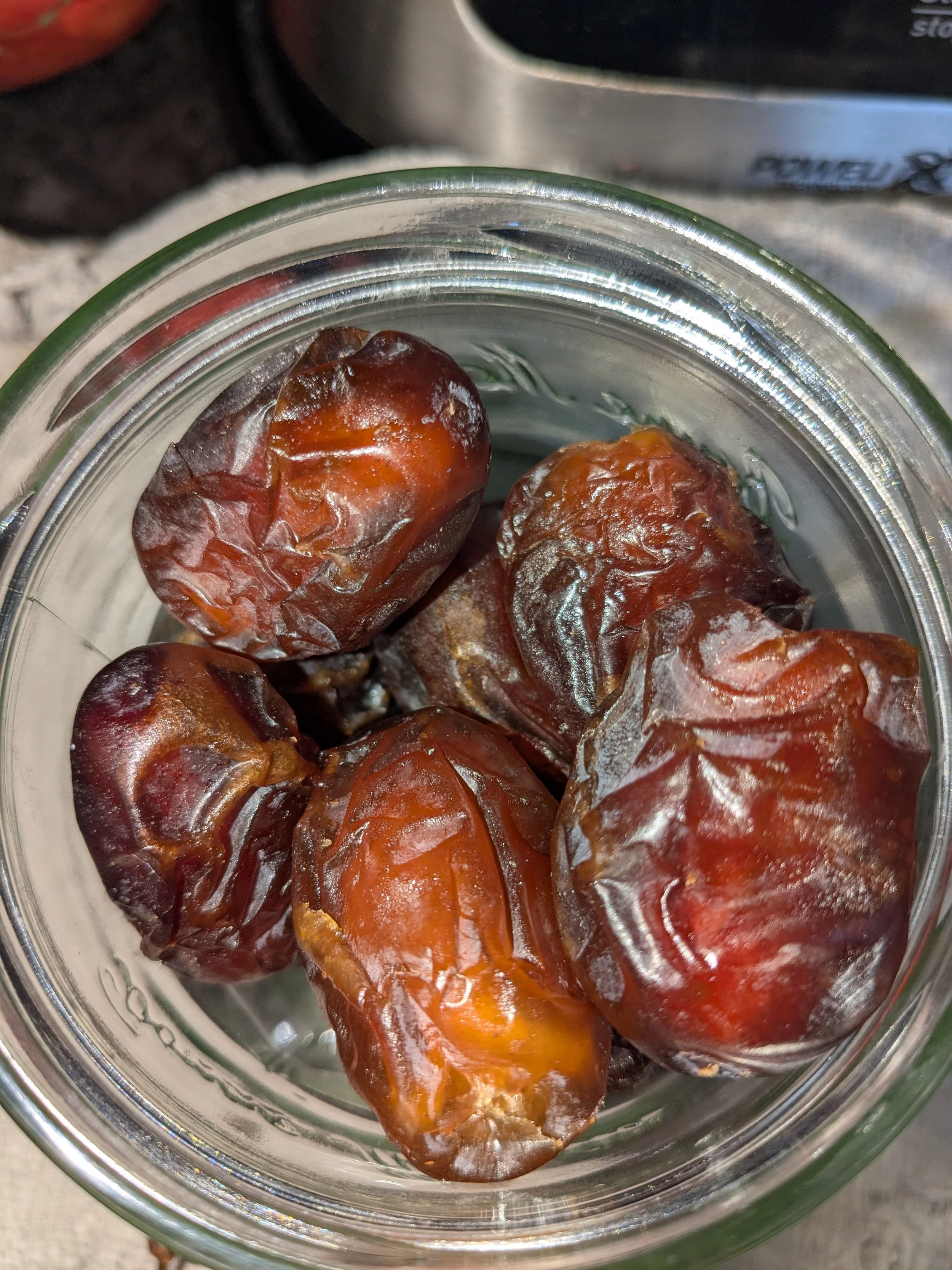 A close-up view of several dates inside a glass jar, with a metal container visible in the background.