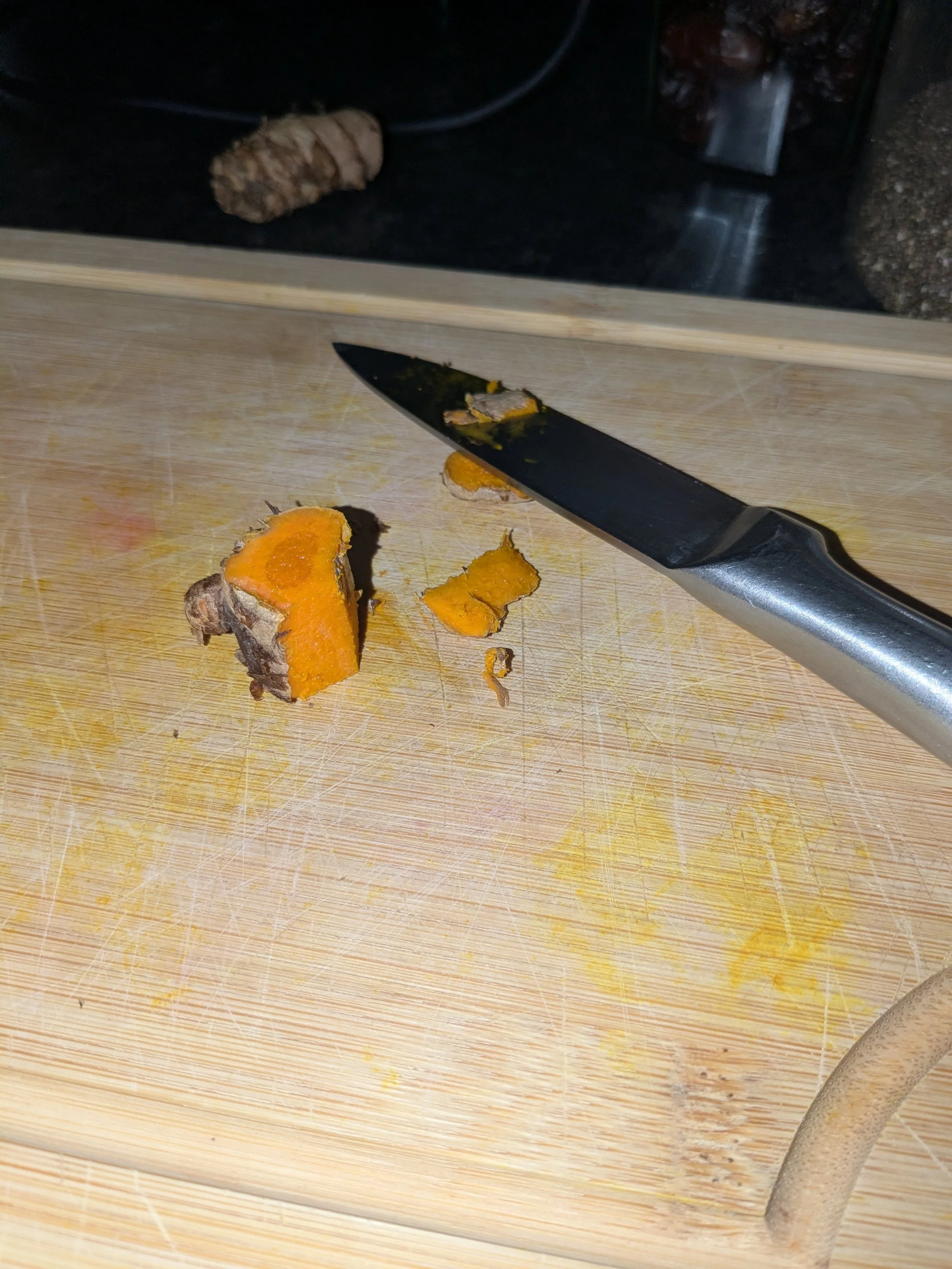 A wooden cutting board with slices of cooked pumpkin and a black knife. In the background, there is a black countertop with a piece of ginger, a clear container with dark round objects, and a packet of food item.