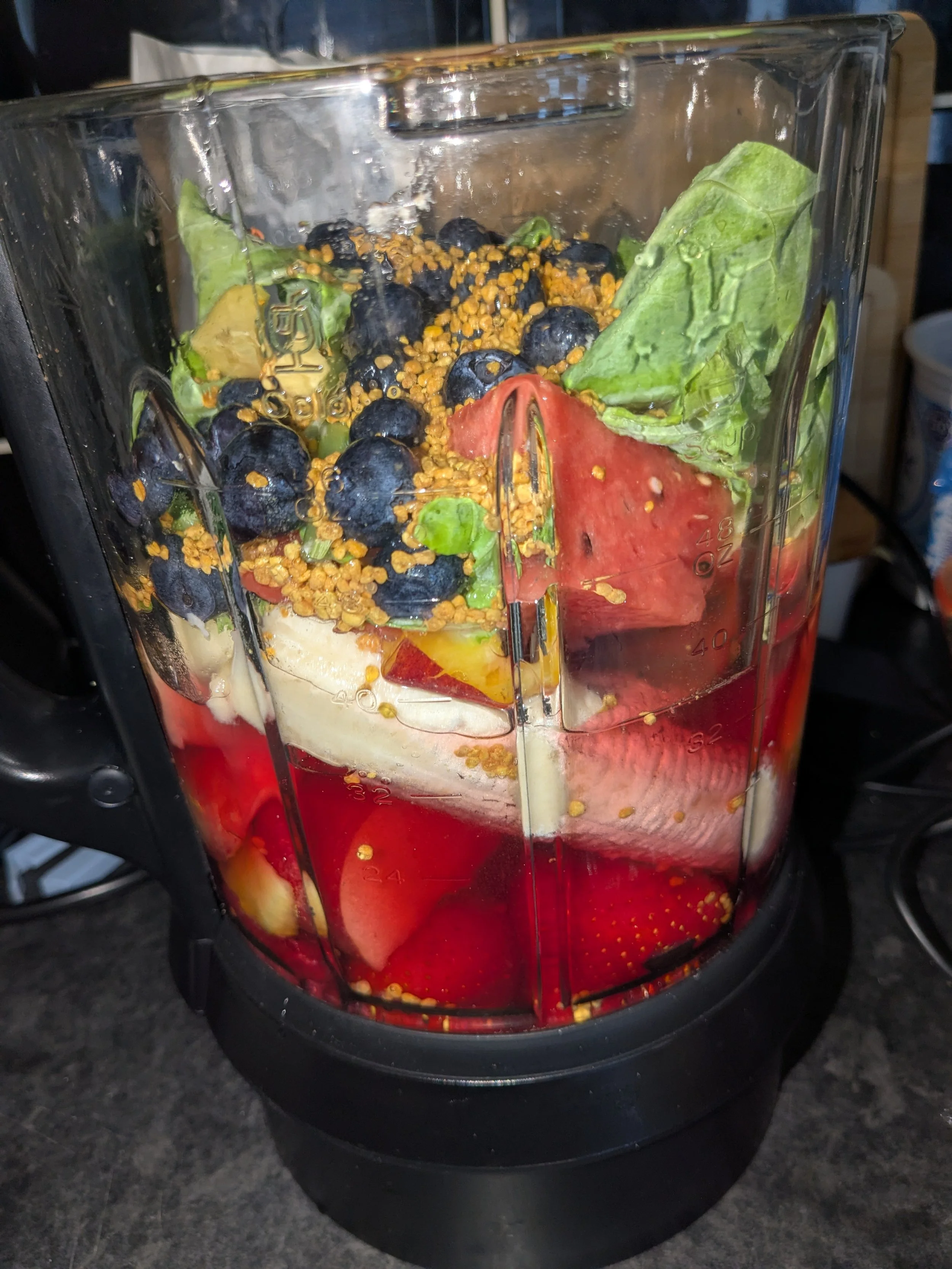 Blender filled with blueberries, banana slices, watermelon chunks, lettuce leaves, and bee pollen.