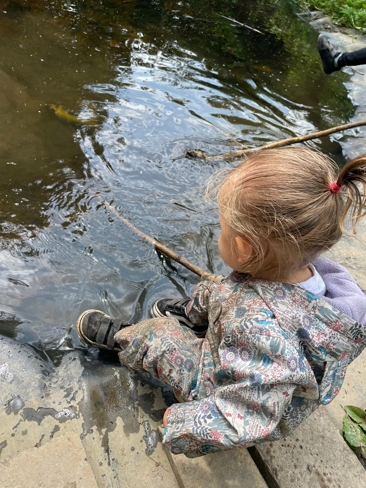 A young girl with a ponytail, wearing a patterned jacket and black shoes, sits by the edge of a pond holding a stick and watching the water.