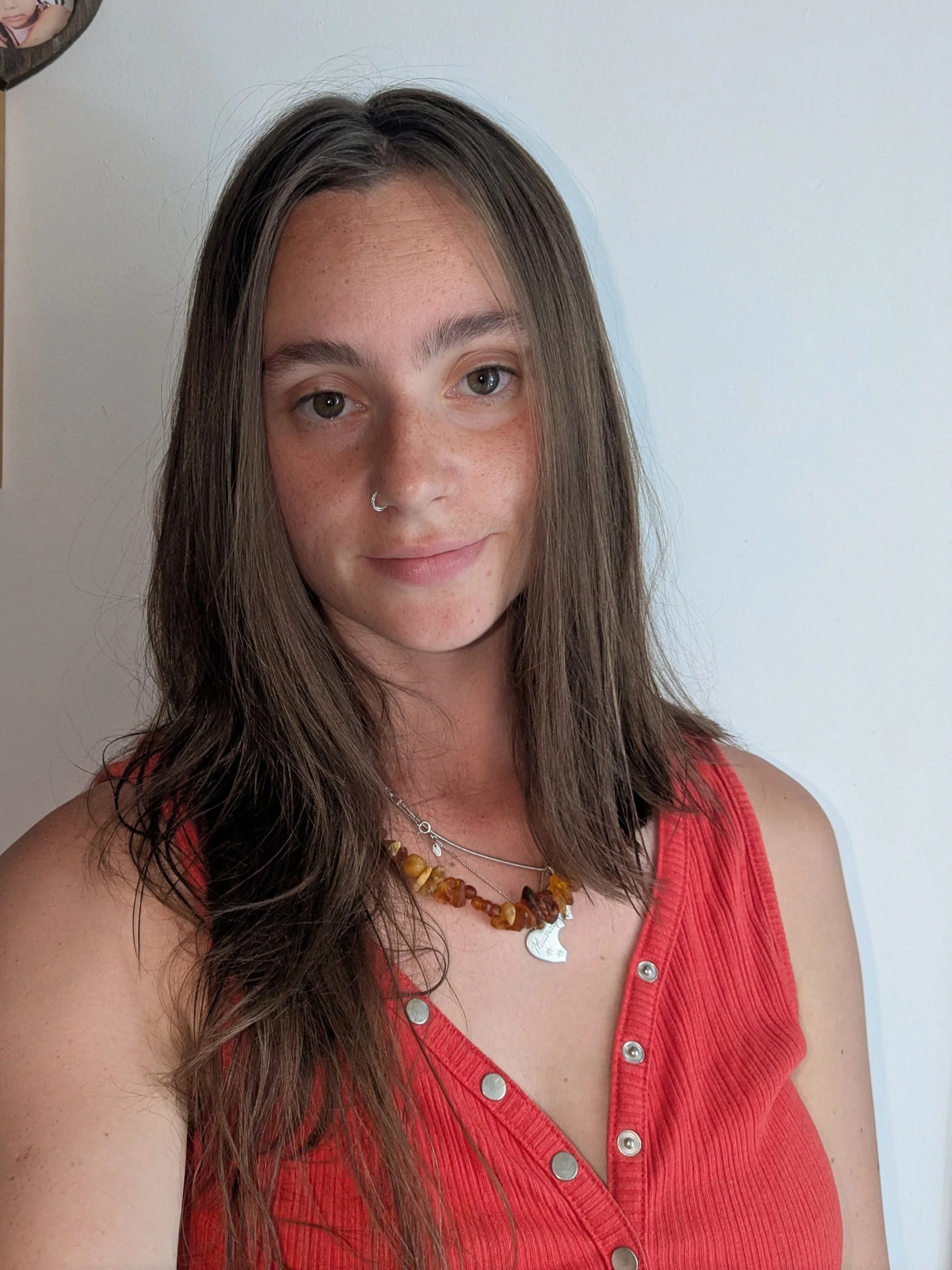 A woman with long brown hair, light skin, and gray eyes, wearing a red sleeveless top with metallic buttons, a silver nose ring, and layered amber-colored necklaces, standing against a white wall.