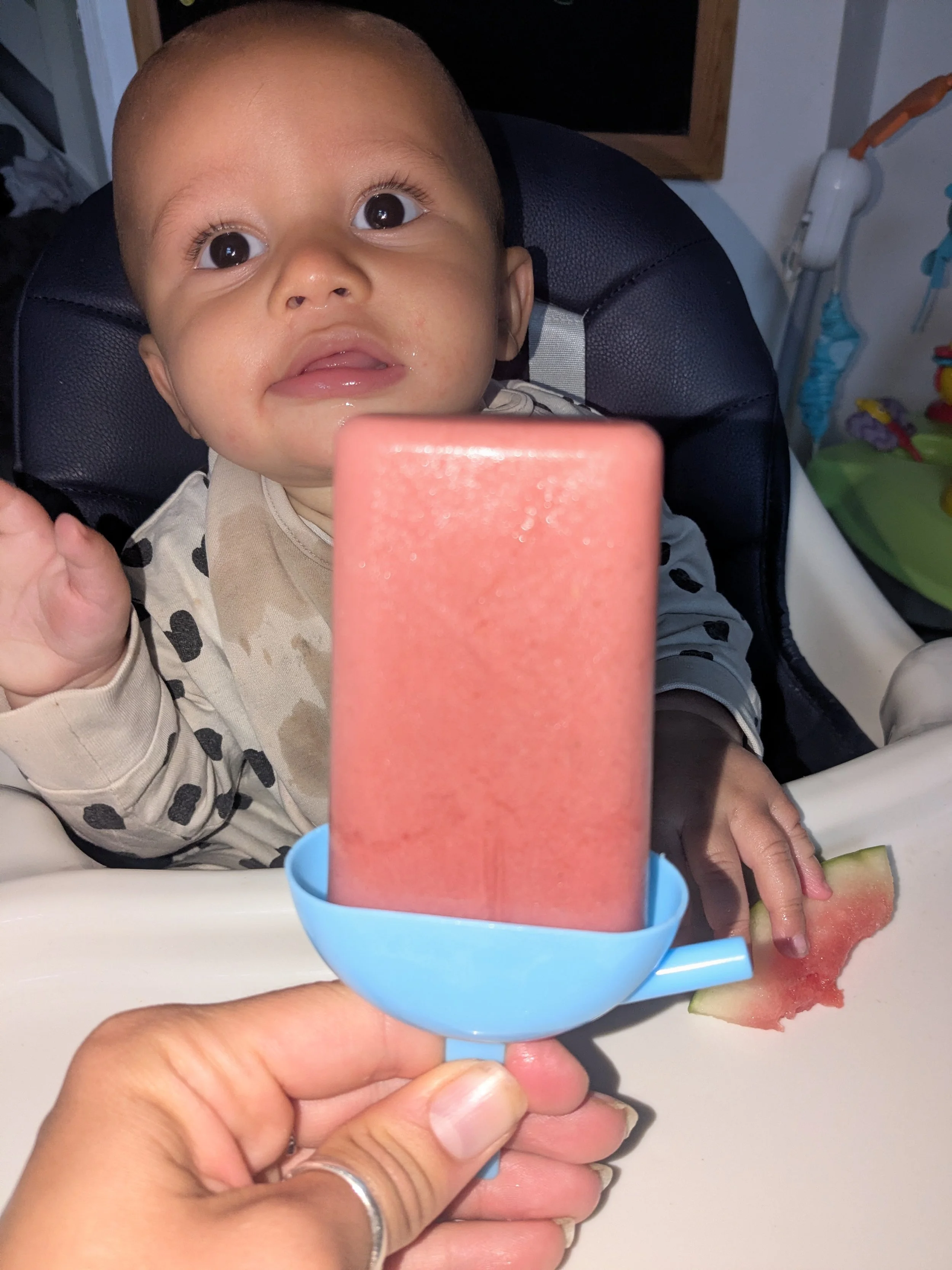 A baby with big eyes is sitting in a high chair, looking up. Someone is holding a pink popsicle in front of the baby, and a piece of watermelon is on the tray in front of the baby.