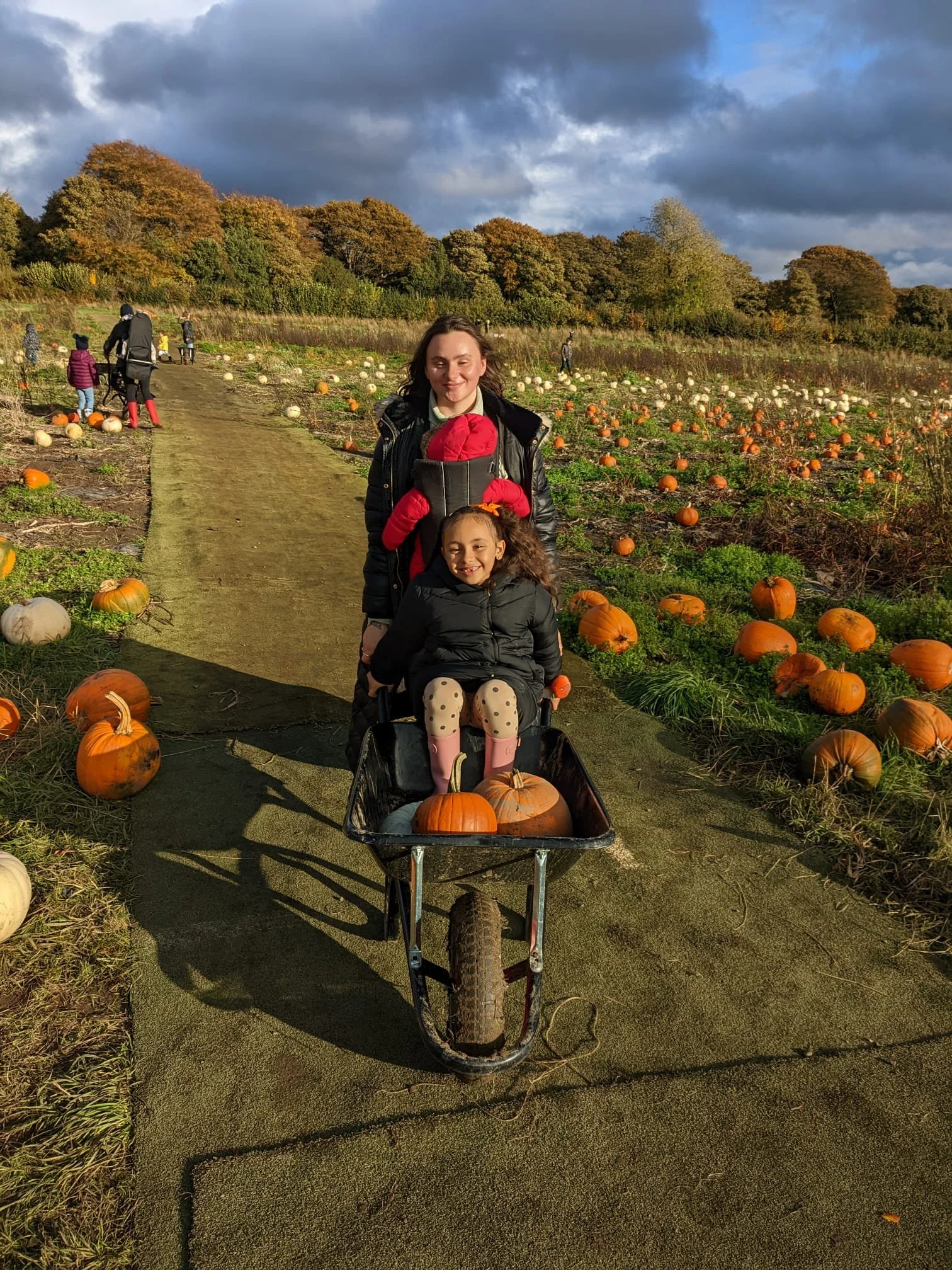 A woman and a girl sitting in a wheelbarrow filled with pumpkins on a pumpkin patch pathway. The woman is smiling with her eyes closed, and the girl has curly hair and is also smiling. Pumpkin patches with scattered pumpkins and other people are visi