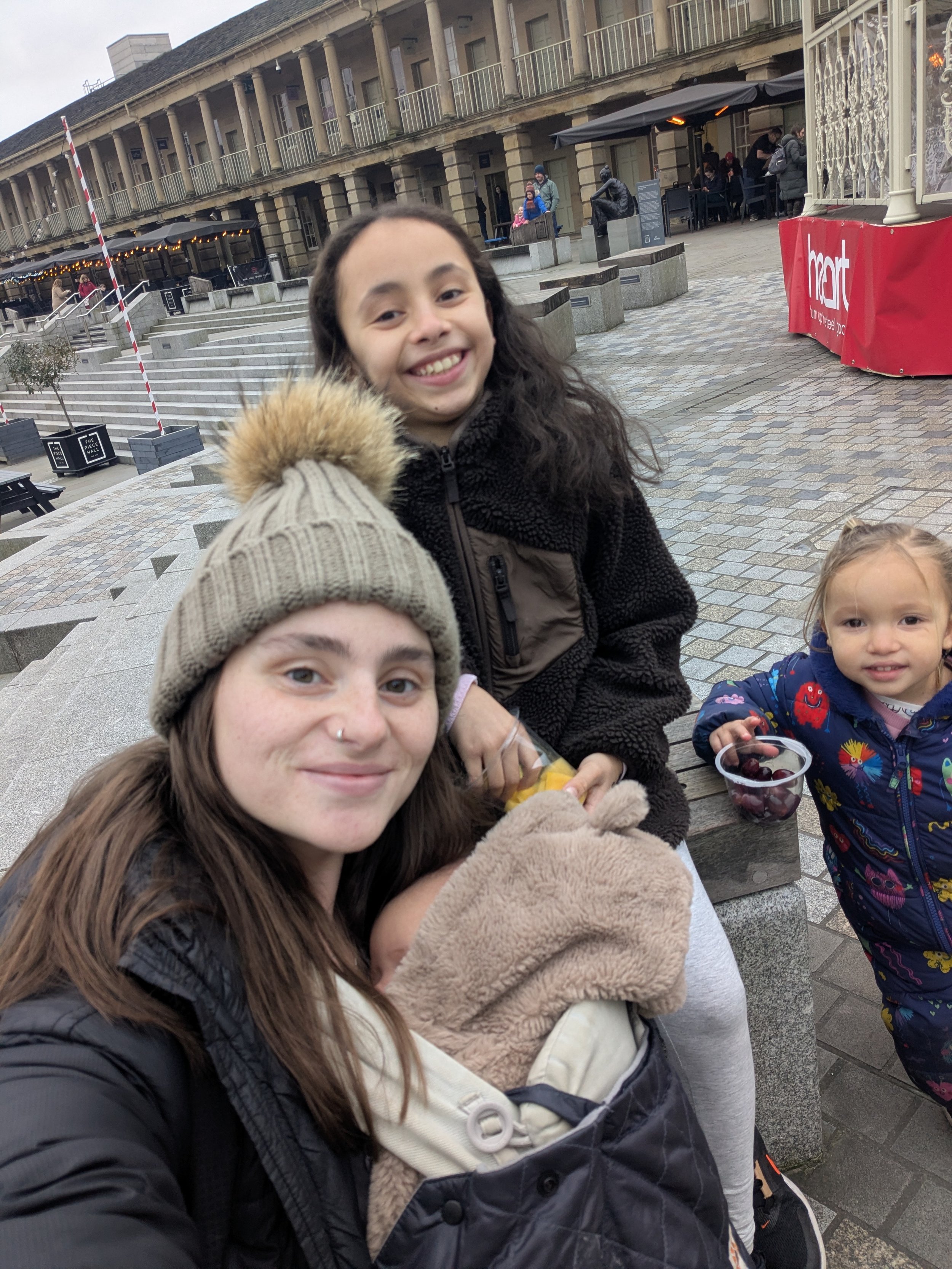 A group of people, including a smiling girl with dark curly hair wearing a black jacket, a person in a beige beanie, and a toddler in a colorful jacket, gathered closely outdoors in a public space with steps, benches, and a building with balconies in the background.