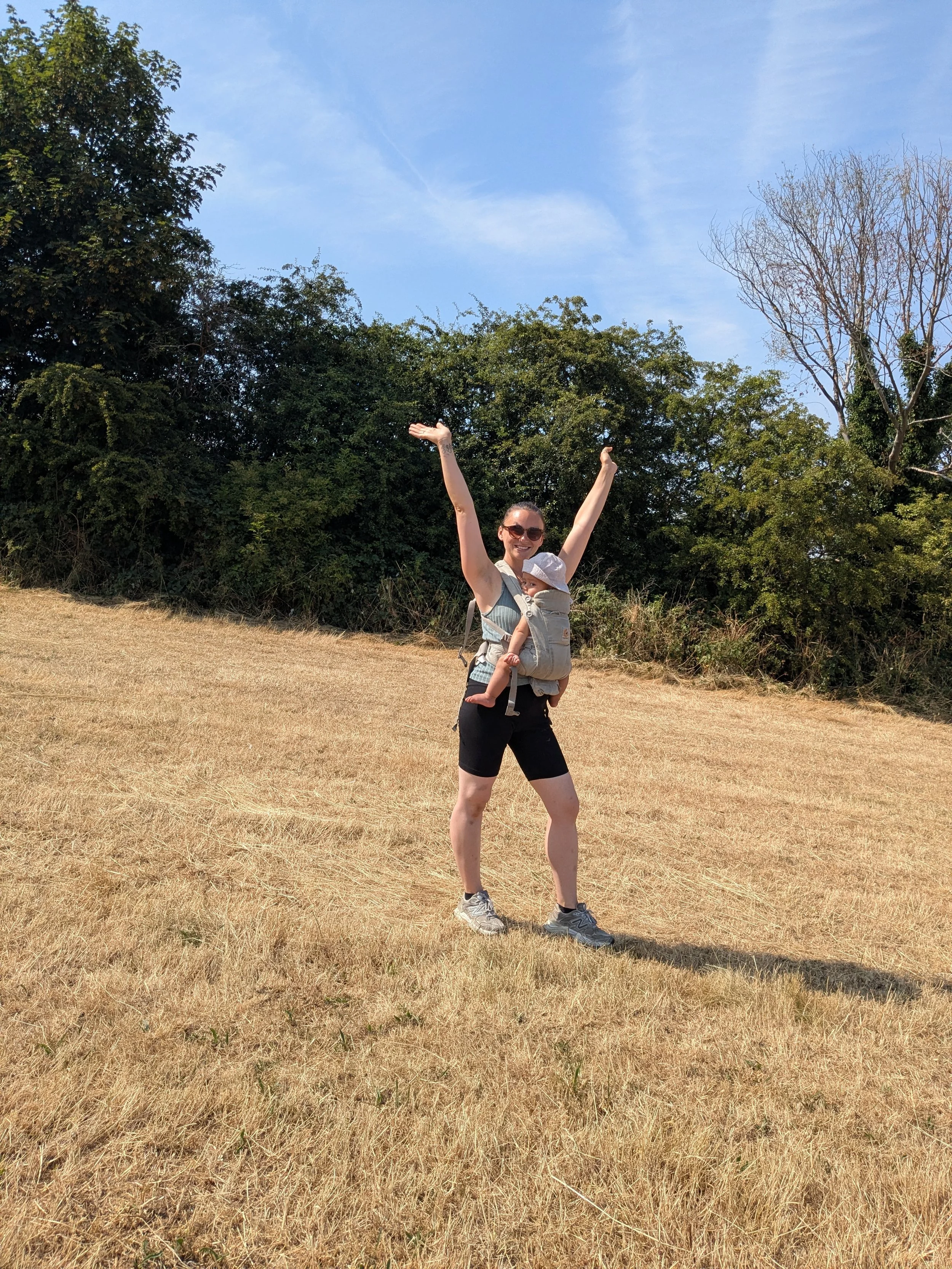 A woman carrying a baby in a baby carrier, standing on a dry grassy field with trees in the background under a blue sky.