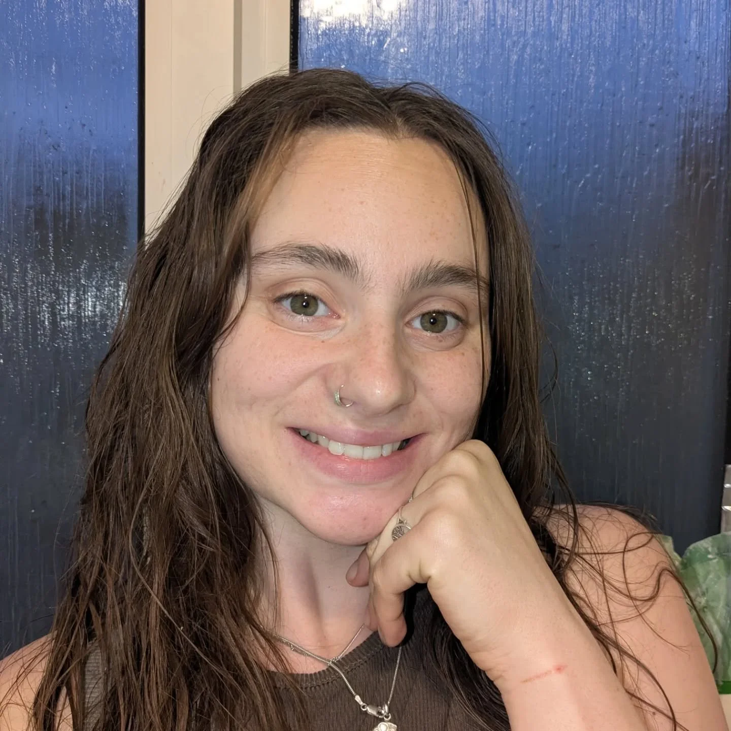 A young woman with long brown hair, light complexion, and light-colored eyes, smiling and resting her chin on her hand. She is wearing a nose ring, a silver necklace, and a ring on her finger. The background features a dark, textured surface.