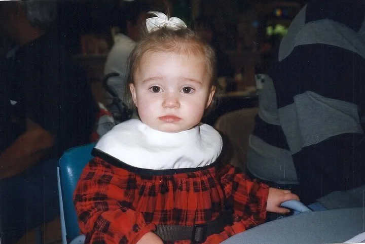 A young girl with light brown hair, wearing a red and black plaid dress and a white bib, sitting at a table in a restaurant or cafe, looking at the camera with a neutral expression.
