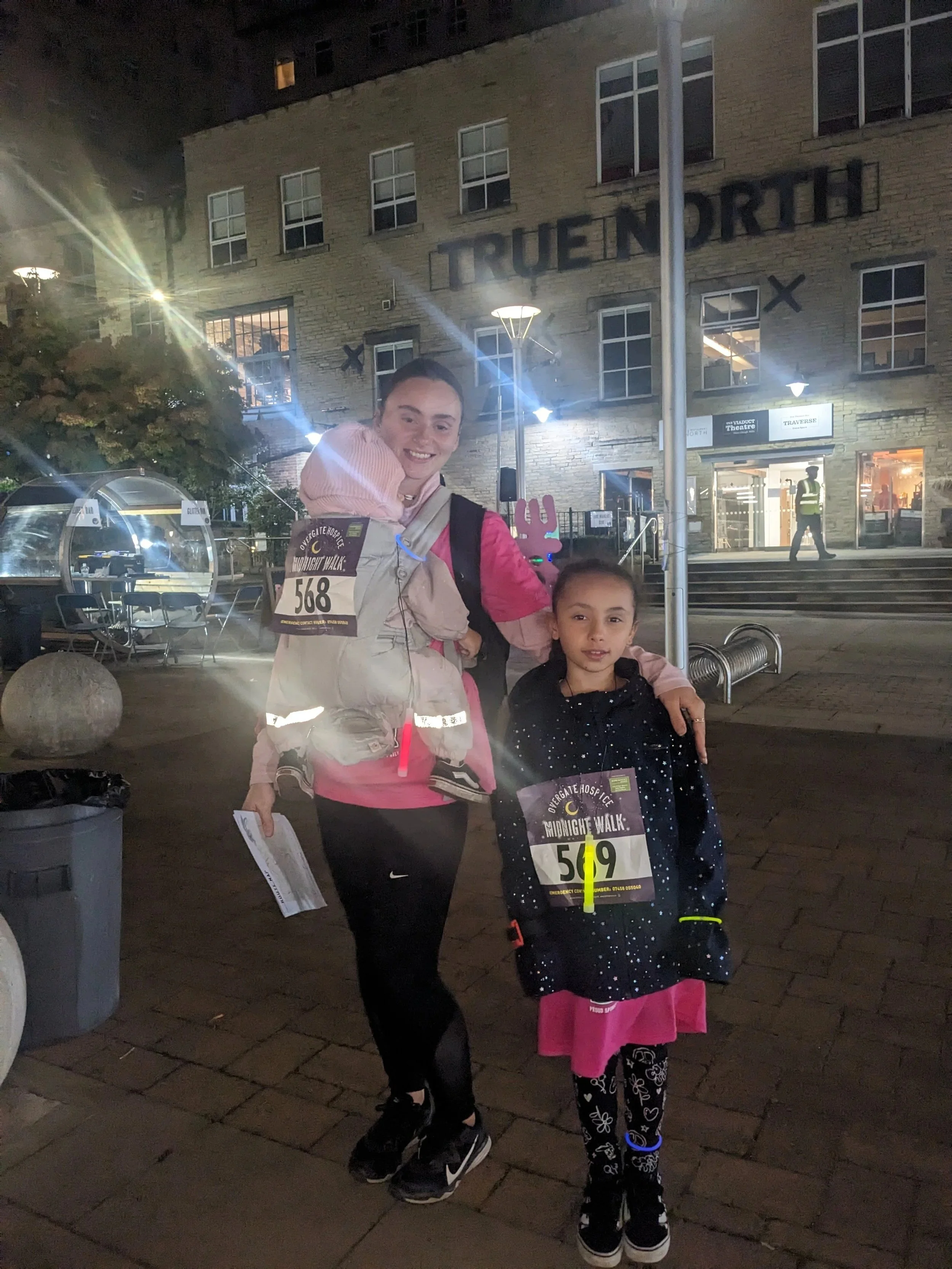 A woman and a young girl participating in a nighttime charity walk outside a building with the sign 'TRUENORTH.' The woman is smiling, wearing a race bib numbered 568, and carrying a child on her back. The girl is holding papers and is wearing a race bib numbered 519.
