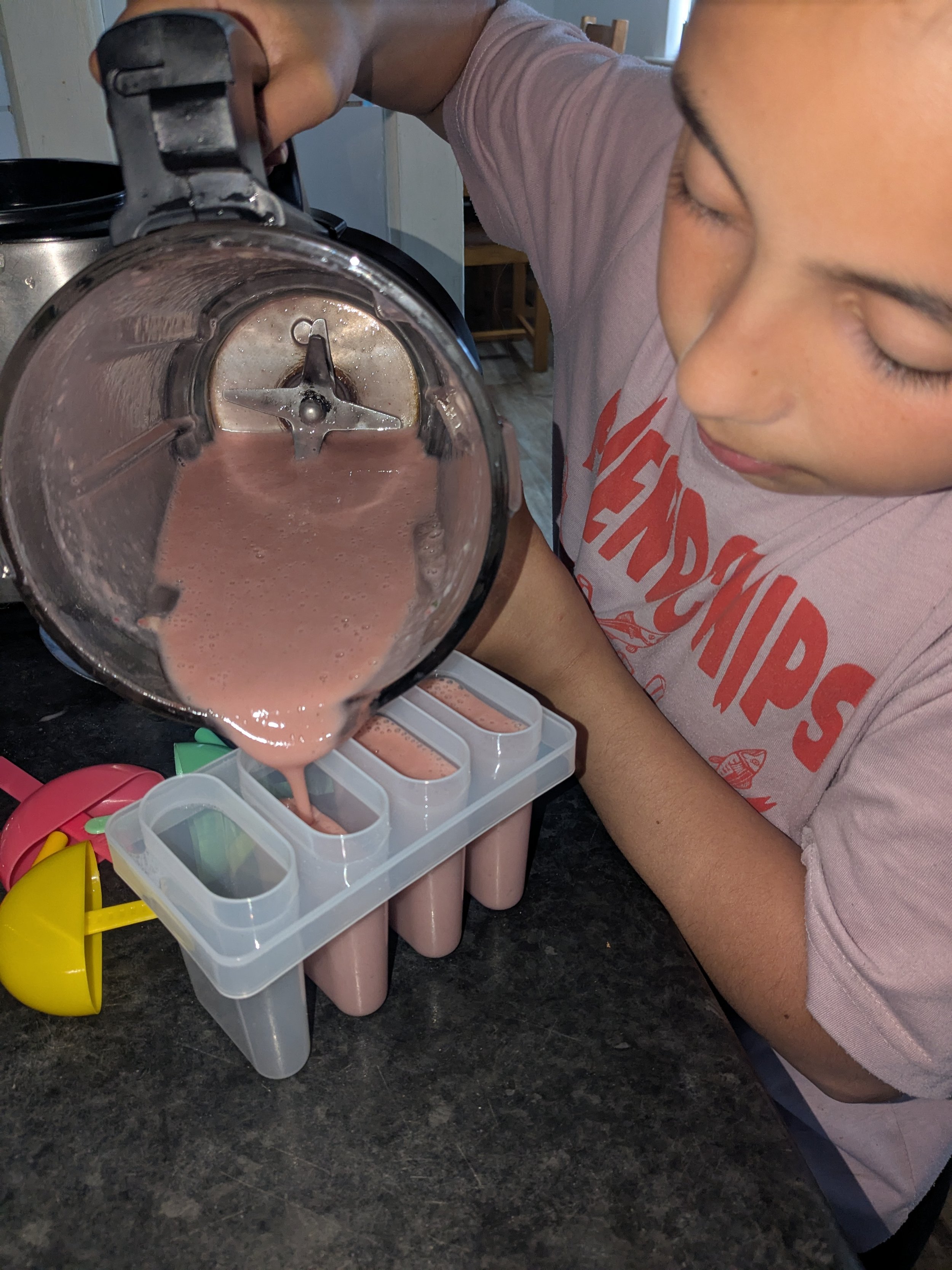 A person pouring a pink smoothie into ice pop molds on a kitchen counter. The person is wearing a light pink t-shirt with red lettering. There are colorful plastic utensils and a yellow bowl nearby.
