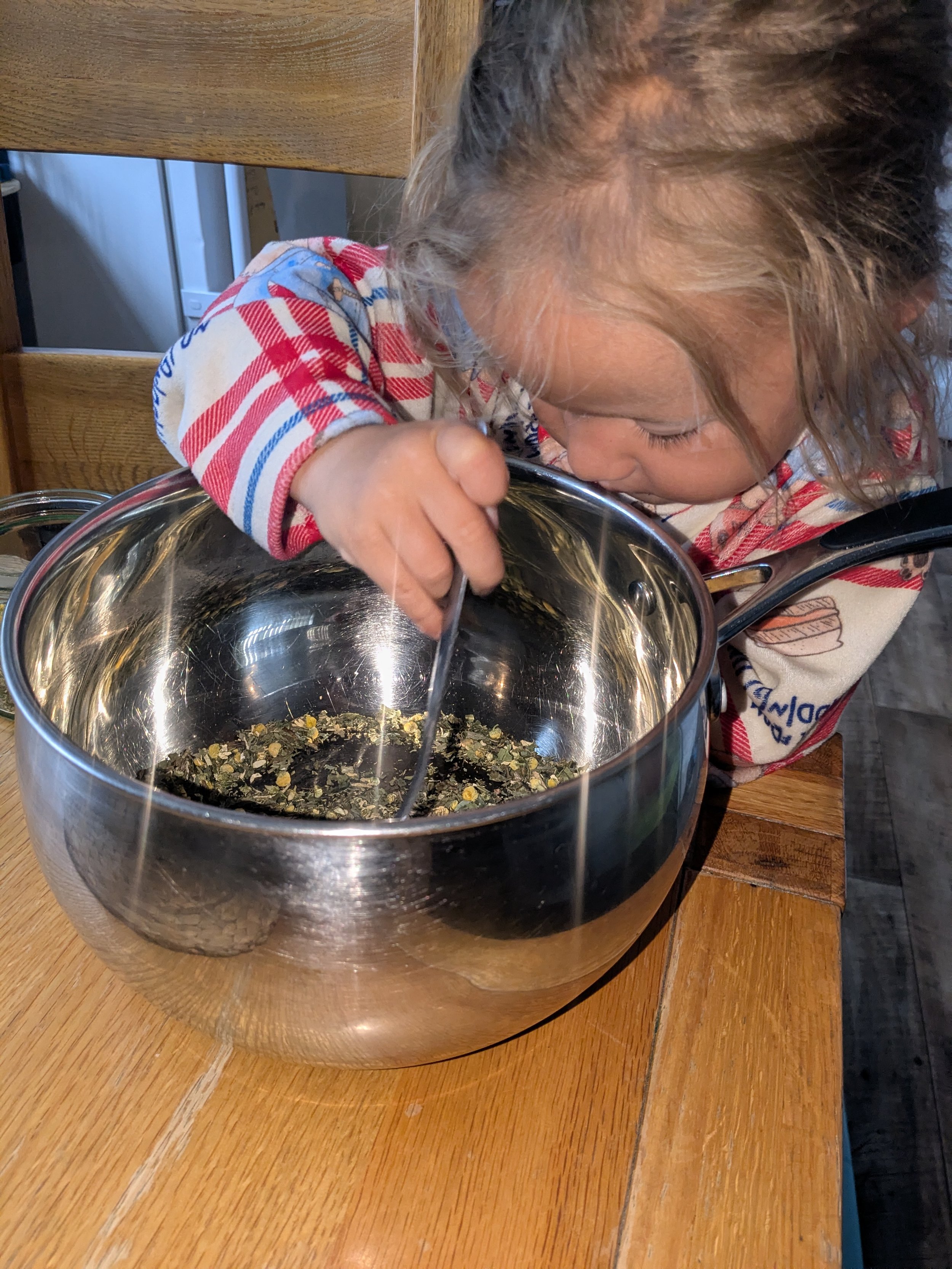 A young girl with curly hair leaning over a large stainless steel mixing bowl with dried herbs and spices, stirring with a spoon, at a wooden table.