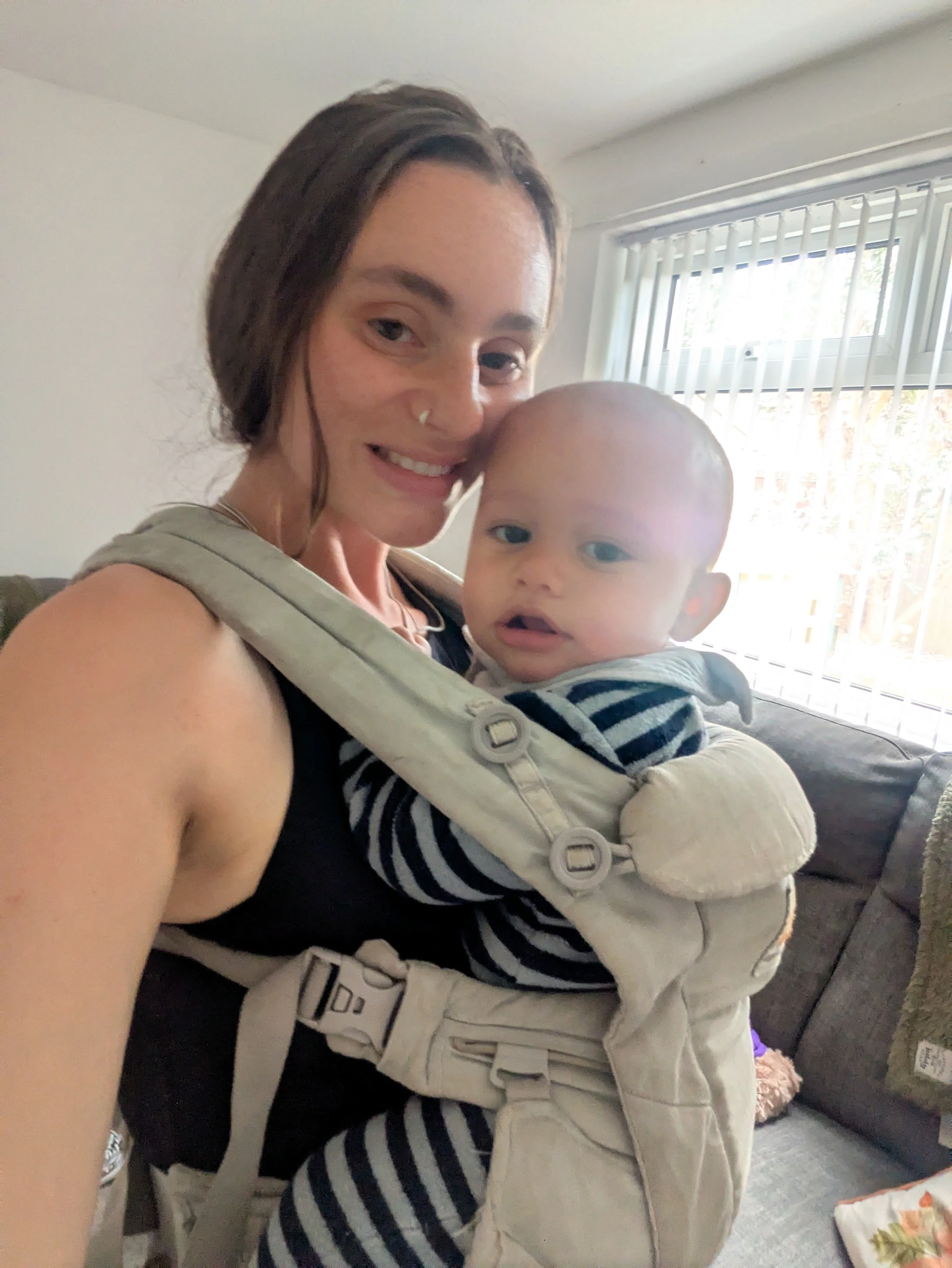 A woman holding a baby with a backpack carrier is smiling in a room with a window and vertical blinds.