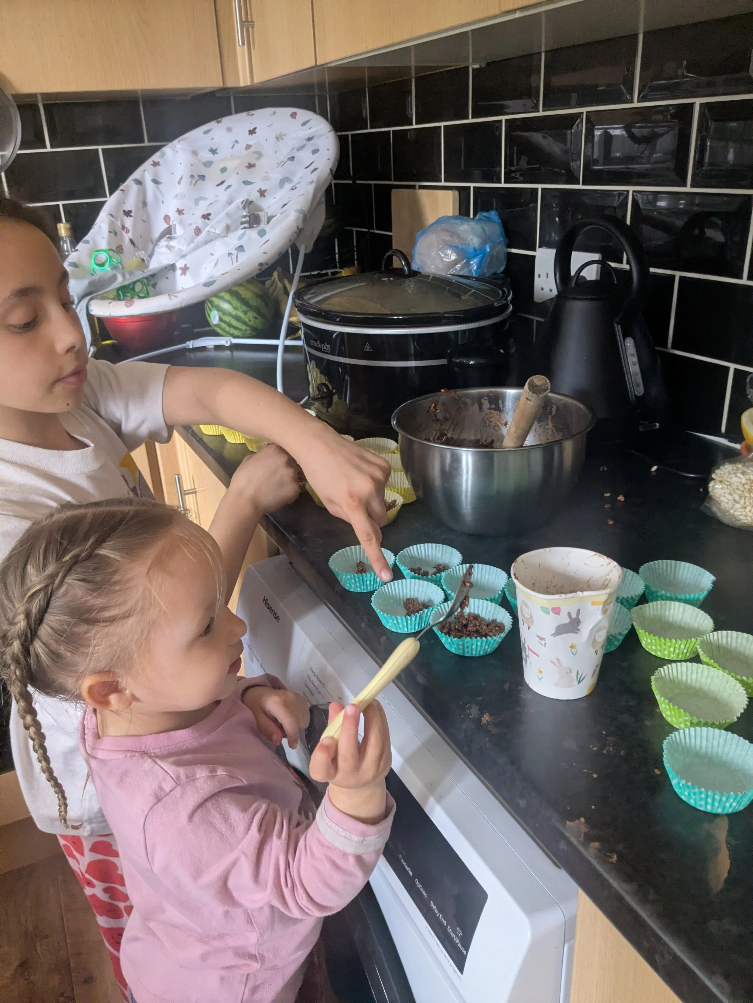 Two young girls are making chocolate cupcakes in a kitchen. One girl is wearing a pink long-sleeve shirt, and the other girl is wearing a white shirt with leopard-print pants. They are filling cupcake liners with chocolate mixture. There are cupcake liners in a cup on the counter, and various kitchen items including a slow cooker, electric kettle, and a plastic bag are visible in the background.
