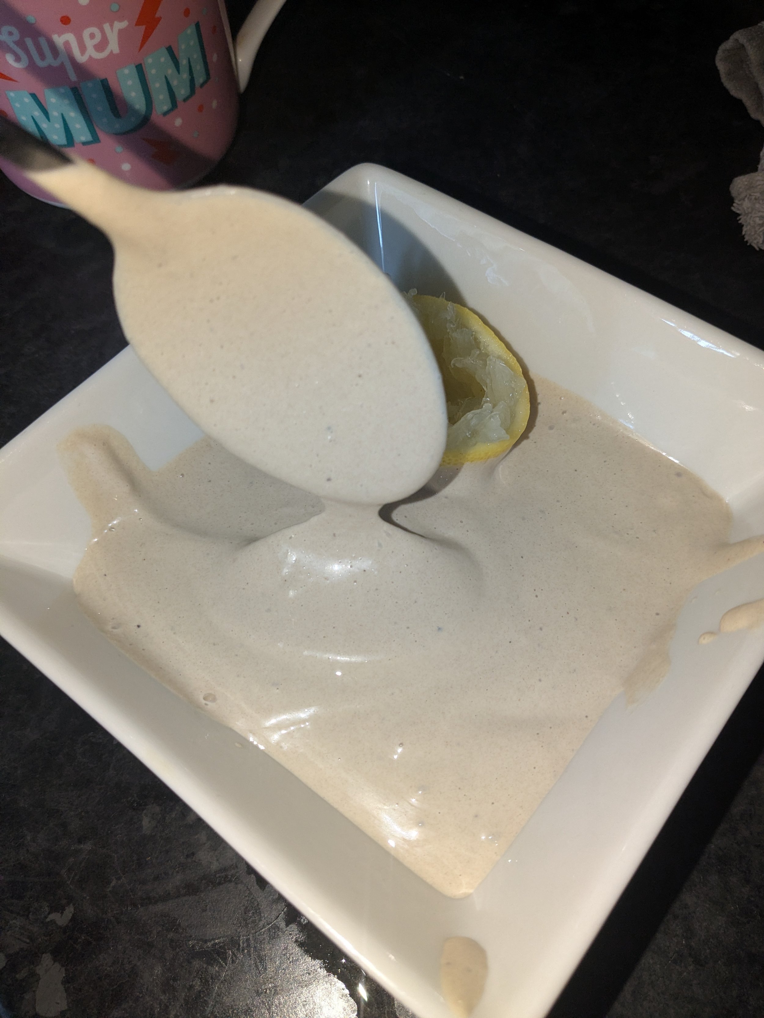 Lemon slice and a spoon pouring a creamy, frothy substance into a white square bowl on a dark surface.