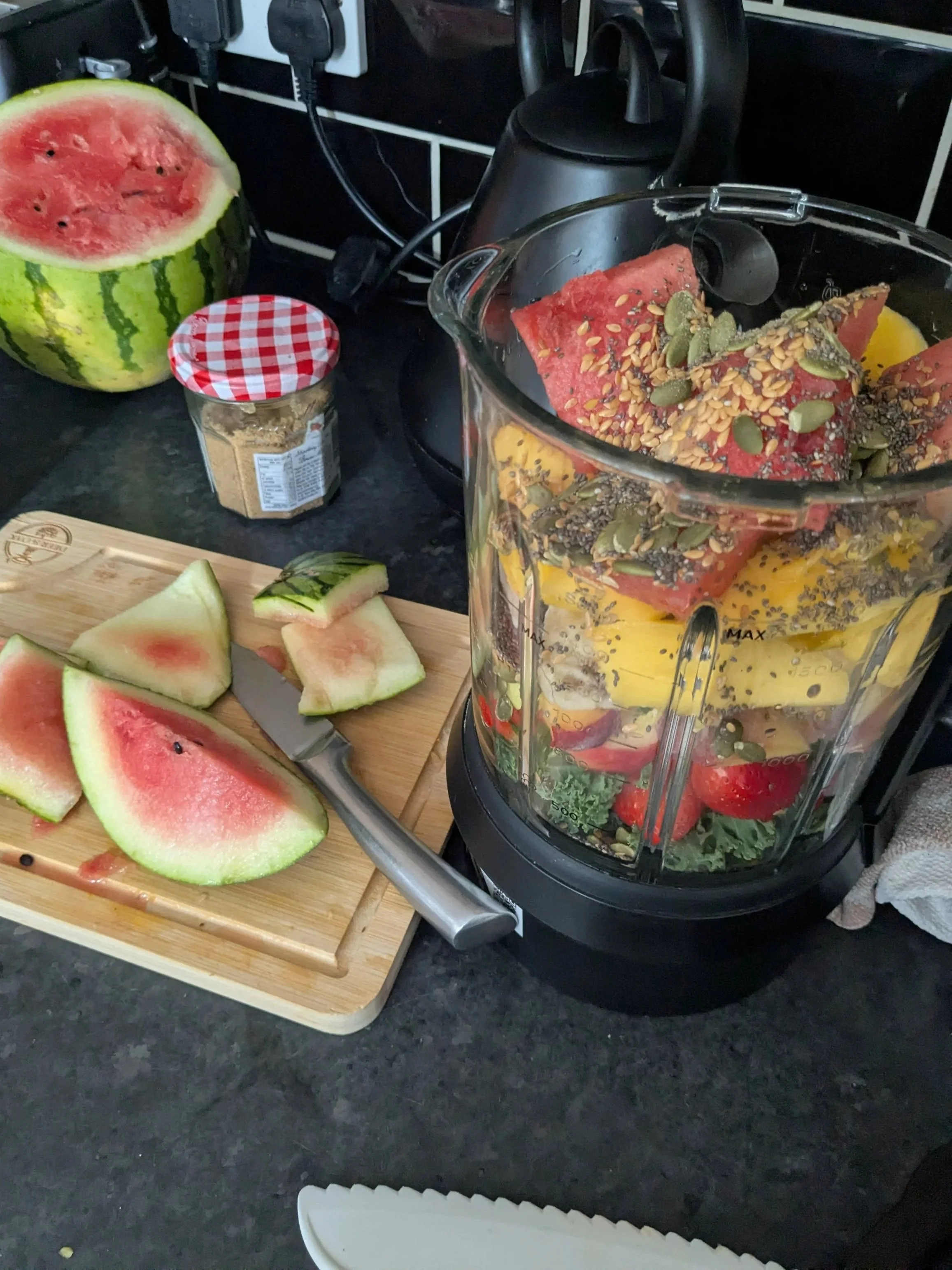 A countertop with sliced watermelon, a jar of honey with a checkered lid, a cutting board, a knife, and a blender filled with various fruits including watermelon, pineapple, and strawberries.