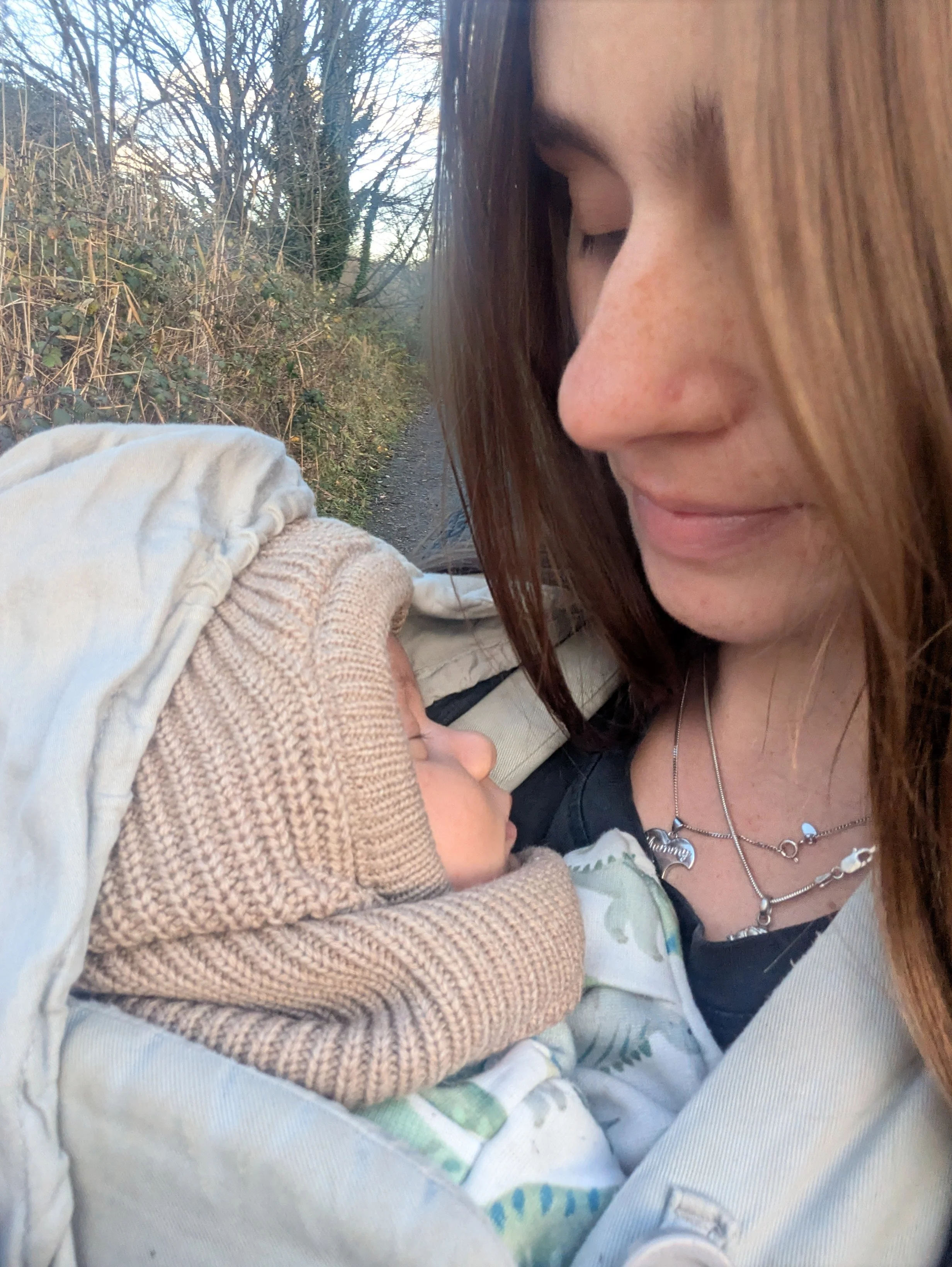 A woman with long brown hair and closed eyes holding a sleeping baby dressed in a beige knit outfit outside on a nature trail with bare trees.