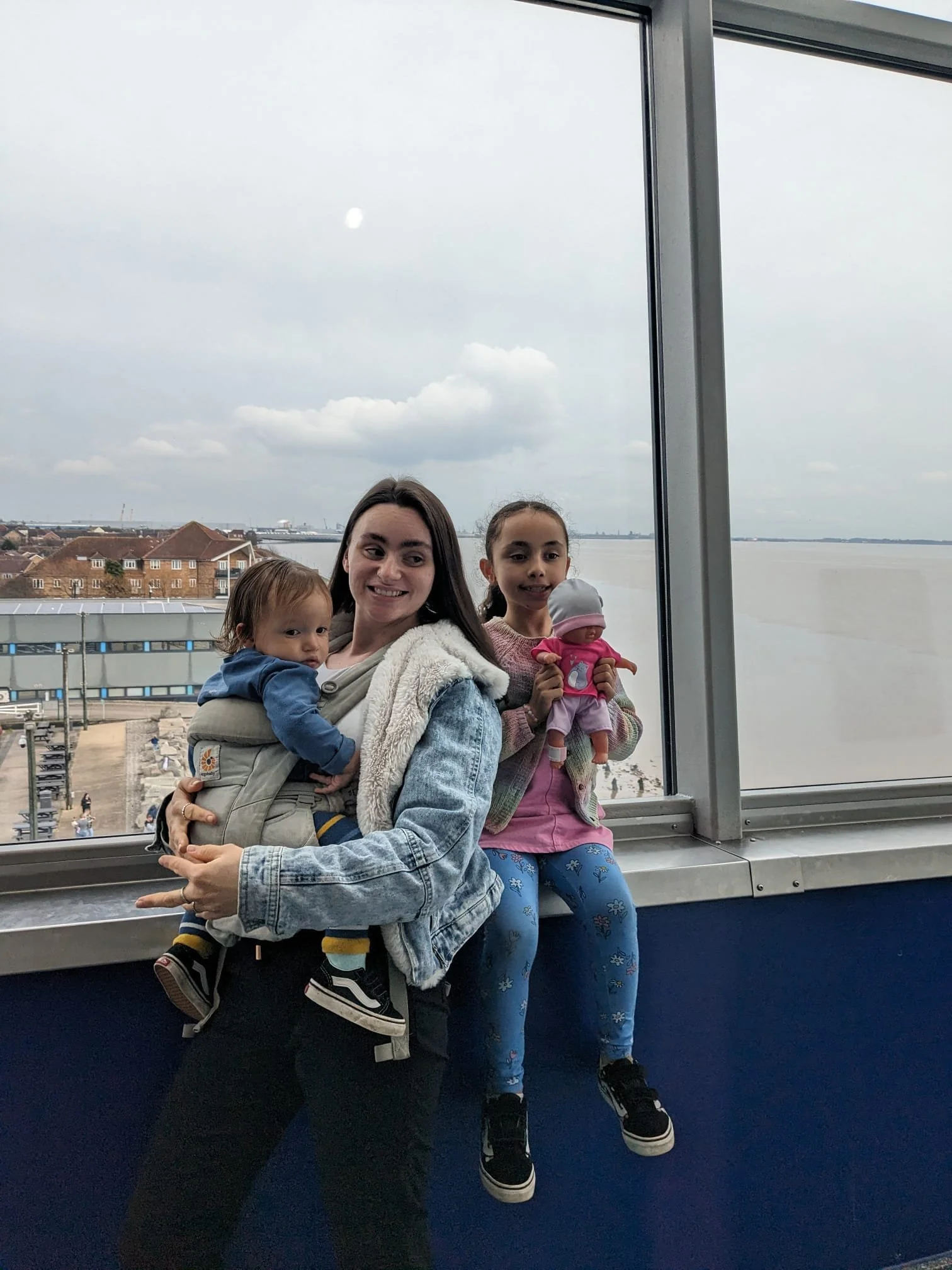 A woman and two young girls sitting on a railing overlooking a waterfront cityscape with cloudy sky.