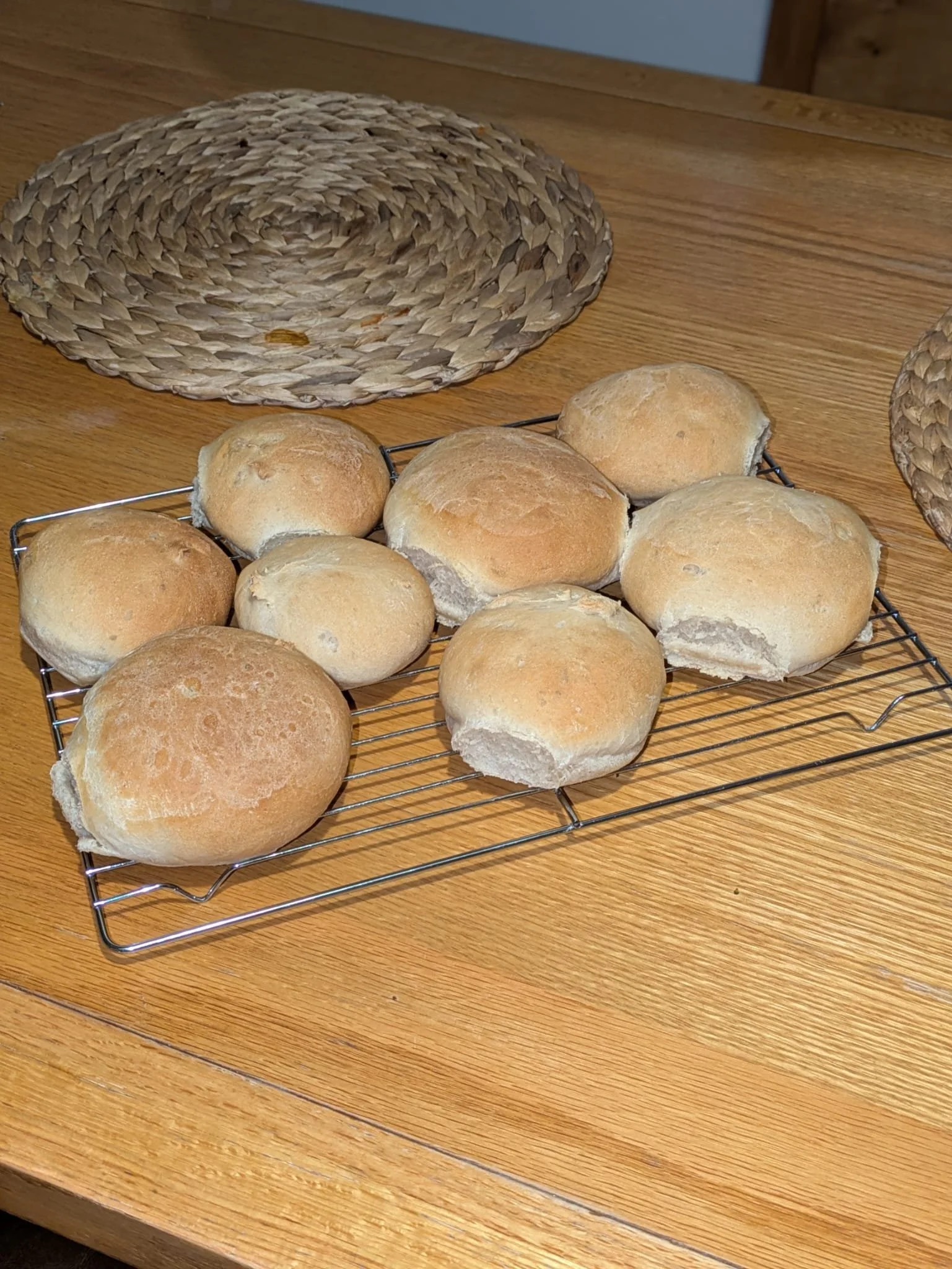 A cooling rack with bread rolls on a wooden table, with a woven placemat and a wooden surface in the background.