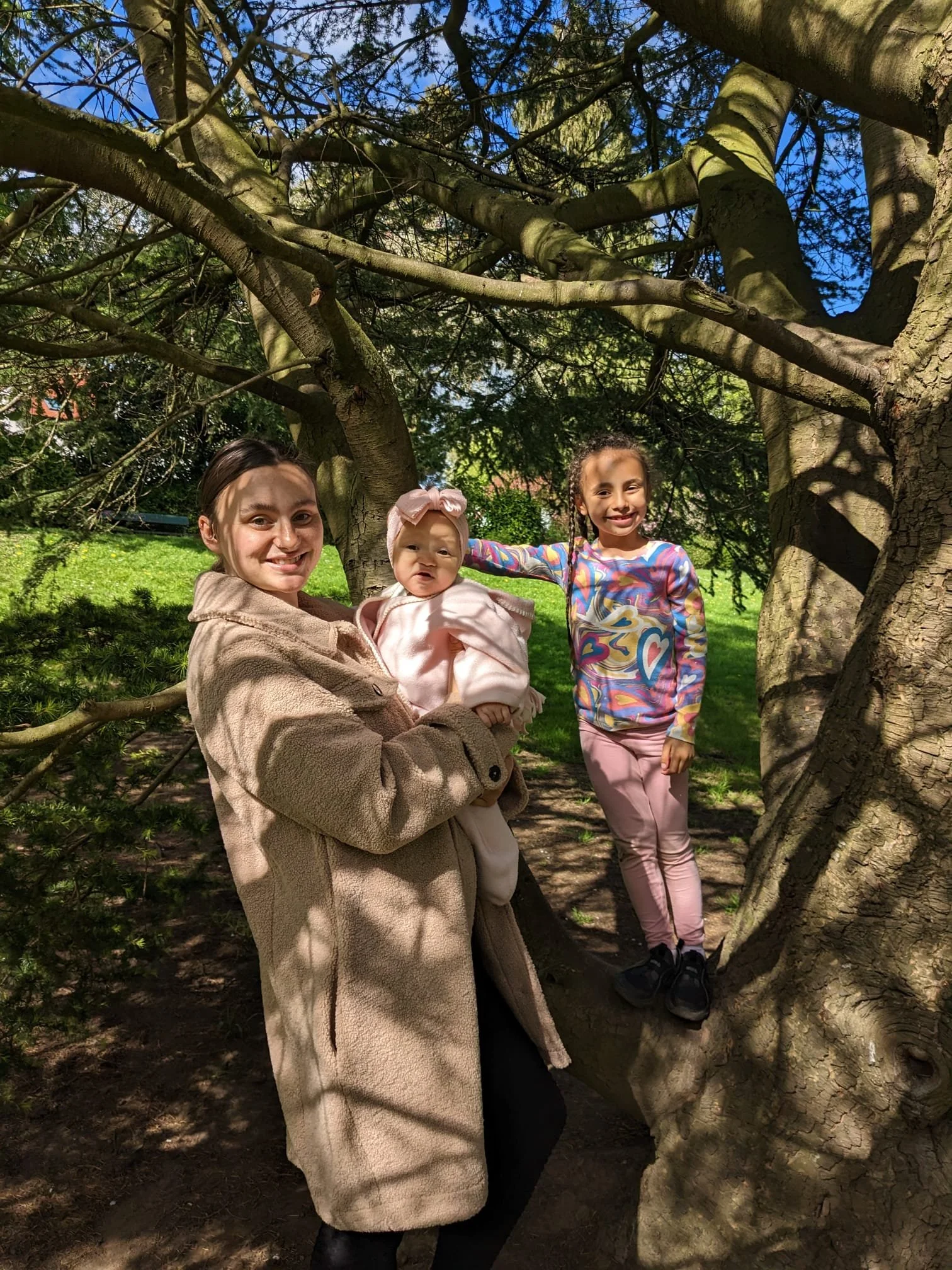 A woman and two young girls near a large tree, smiling and posing outdoors under a blue sky with clouds.