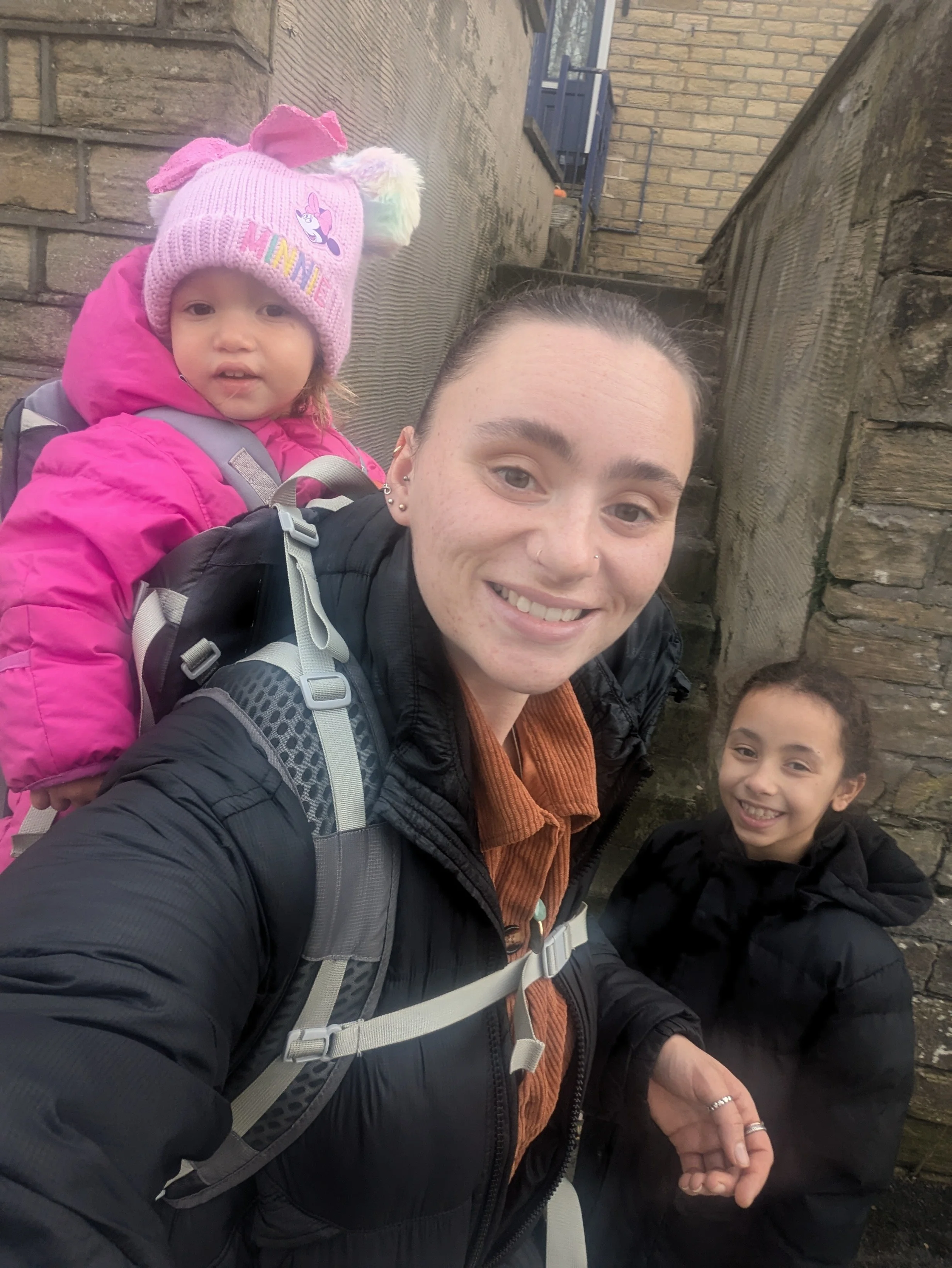 A woman with a backpack smiling while taking a selfie with two children, one girl with a pink jacket and hat with rainbow and unicorn designs, and a boy with a black jacket, standing outdoors against a brick and stone wall.