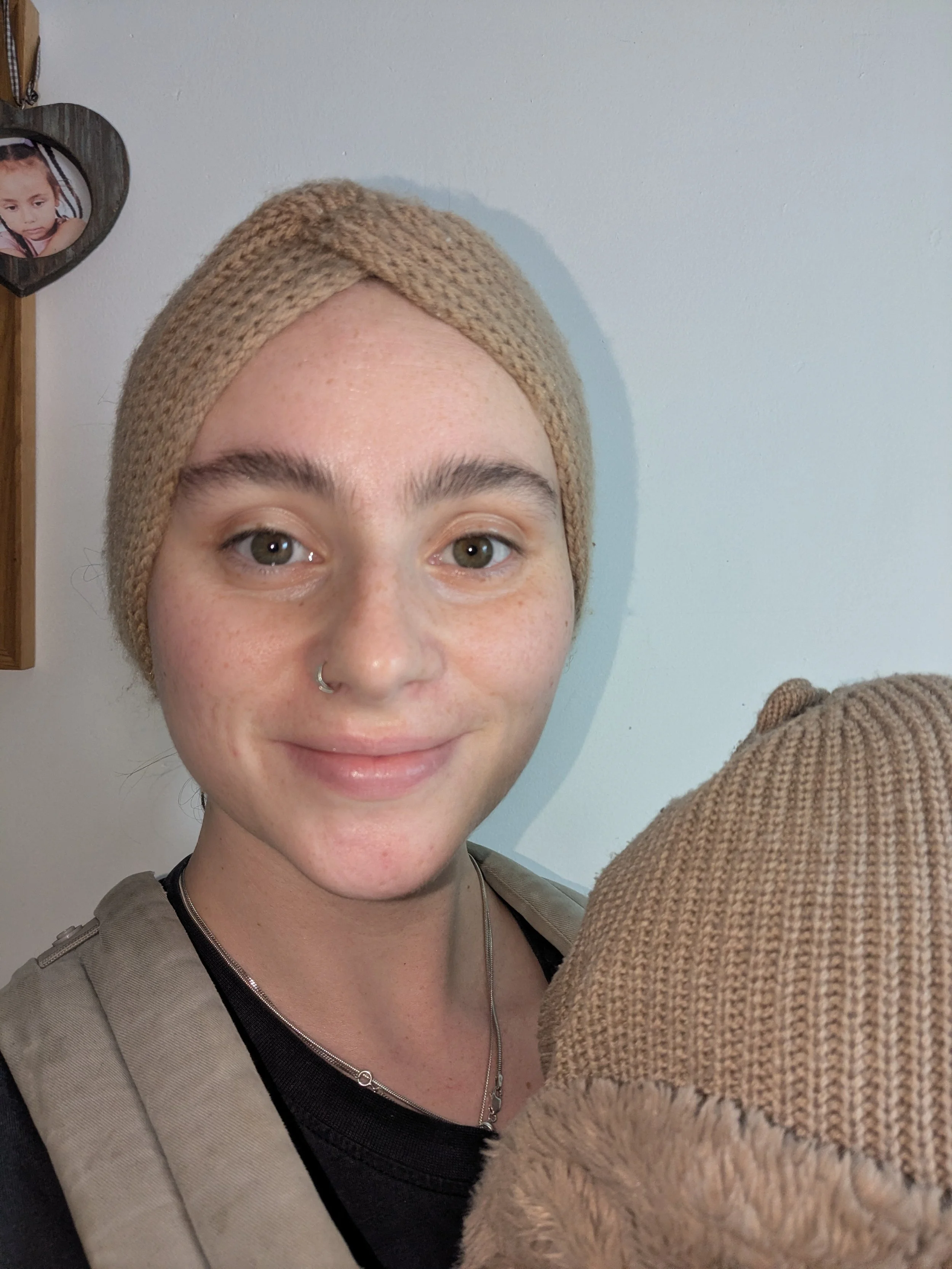 A woman with short hair, light skin, and a nose ring wearing a beige knit headband, gray and black clothing, is smiling while holding a brown knit teddy bear in front of her face. The background is a plain white wall with a photo of a girl inside a h
