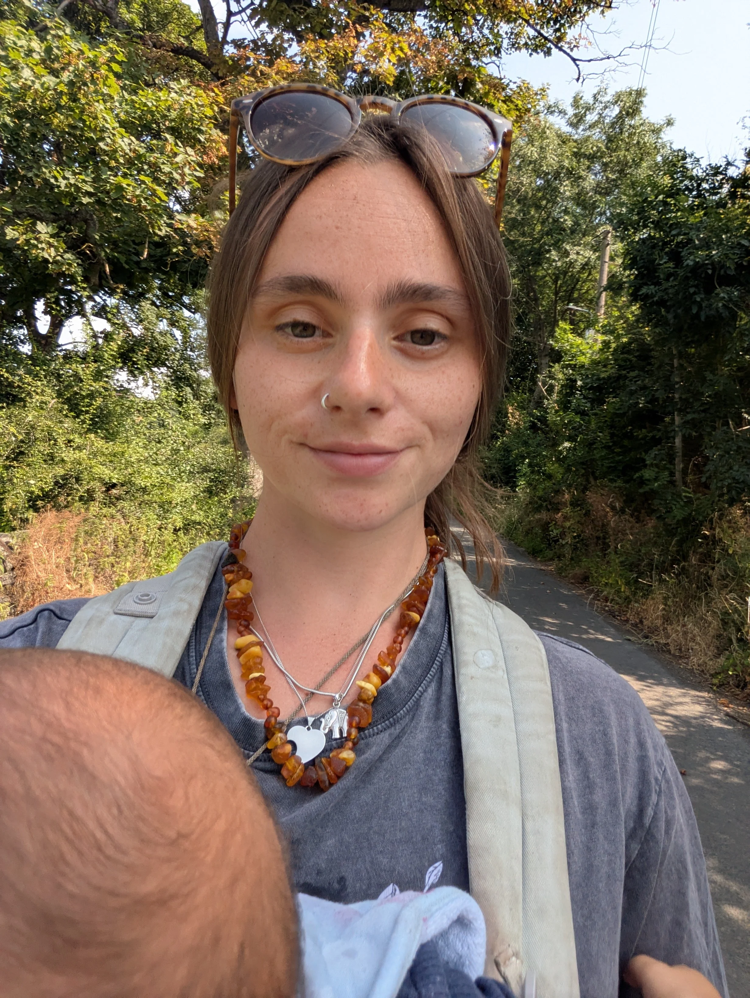 Young woman with sunglasses on her head, wearing necklaces and a dark t-shirt, walking outdoors on a sunny day surrounded by trees and greenery.