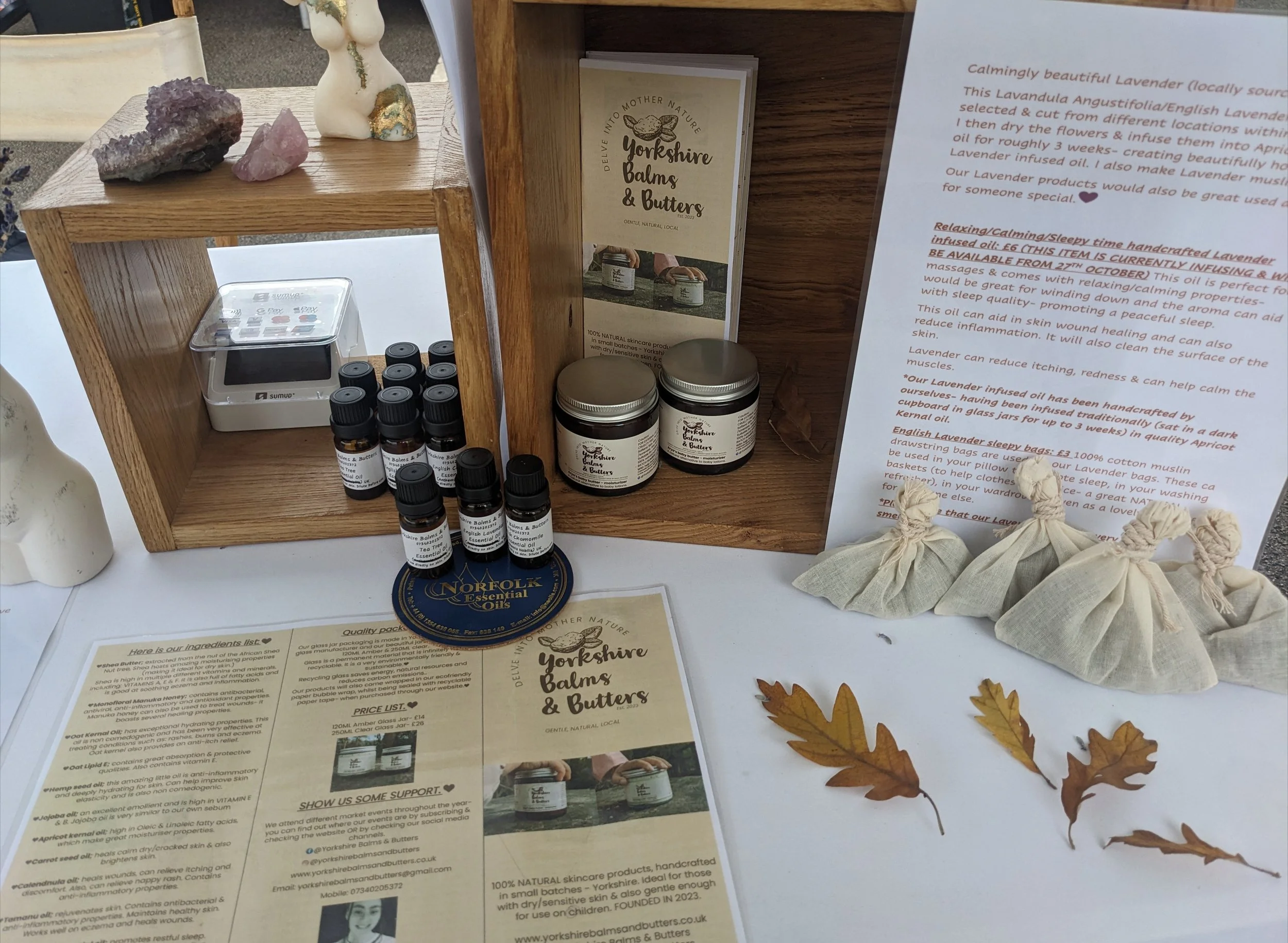 A display at a market stand featuring small jars of lavender balm, a informational leaflet from Yorkshire Balms & Butters, some amethyst and rose quartz crystals, a small bunny figurine, and several dried autumn leaves. The leaflet provides informati