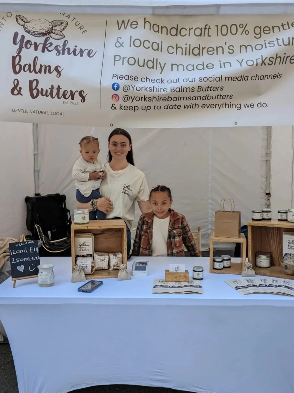 A woman and two children behind a table at a Yorkshire Balms & Butters booth, displaying jars of balms, with a large banner above promoting handcrafted natural skincare products.