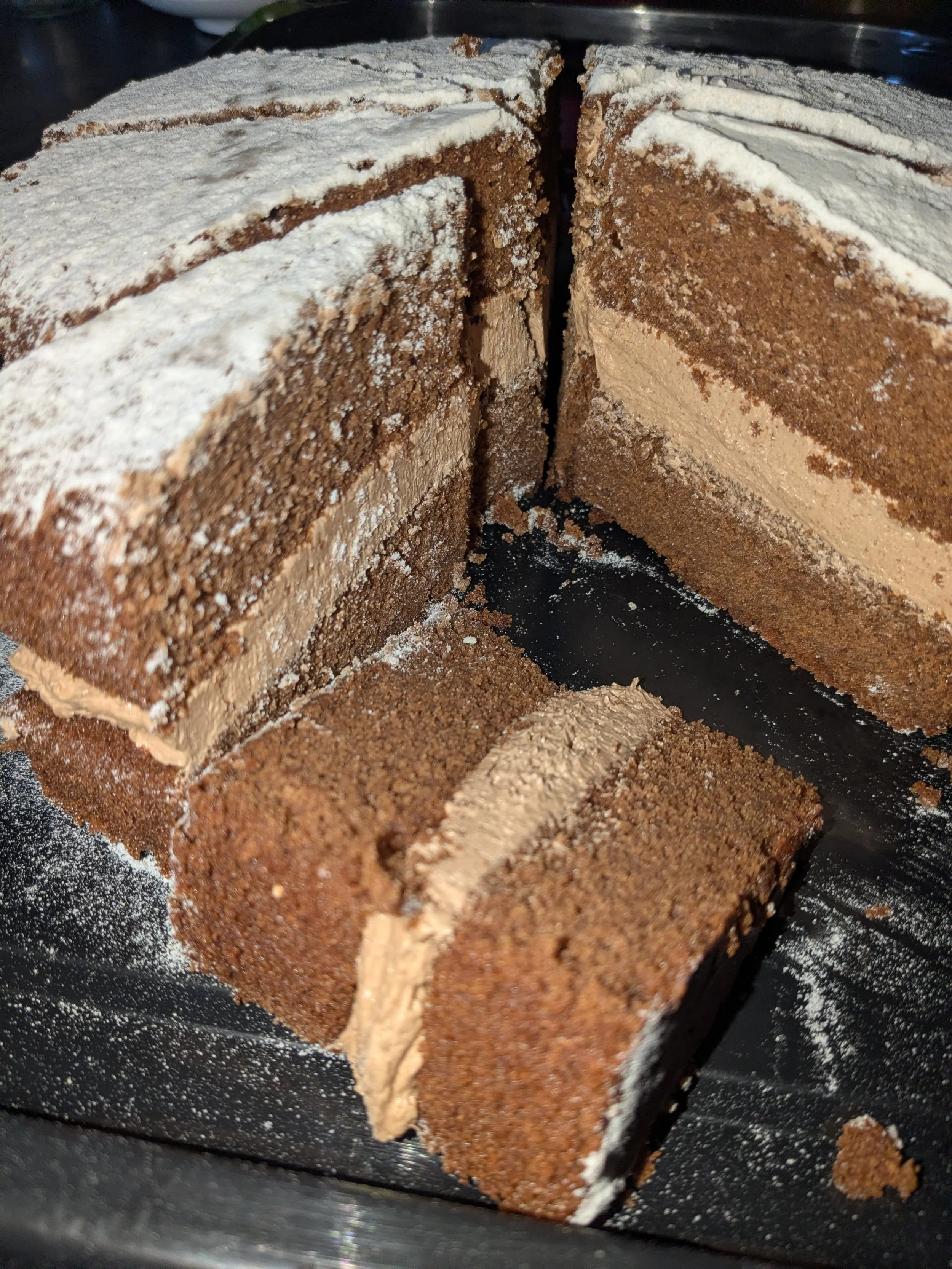 A sliced layered chocolate cake with cream and chocolate powder topping on a black tray.
