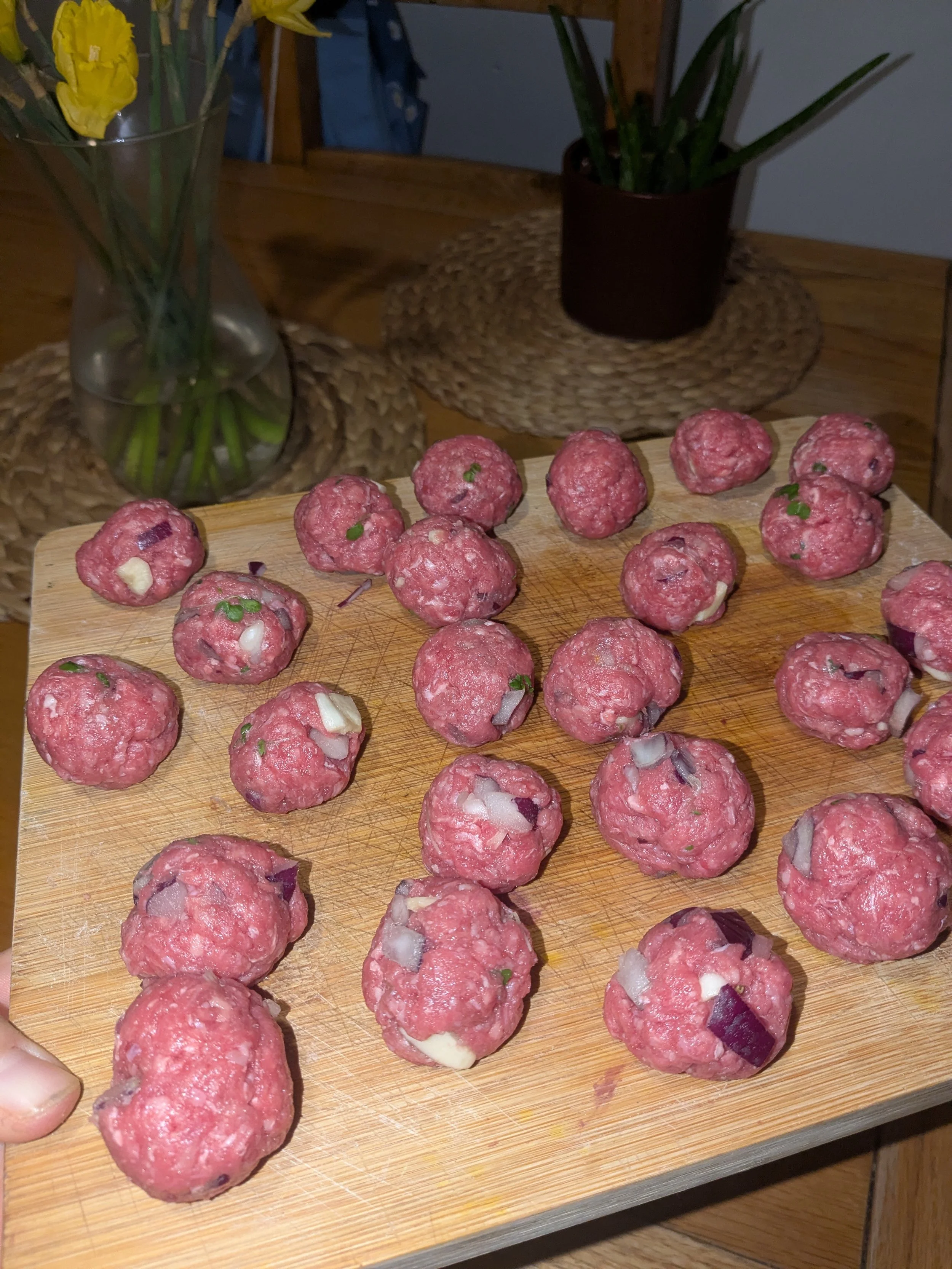 Raw ground beef shaped into small meatballs with visible pieces of onion and green herbs on a wooden cutting board.