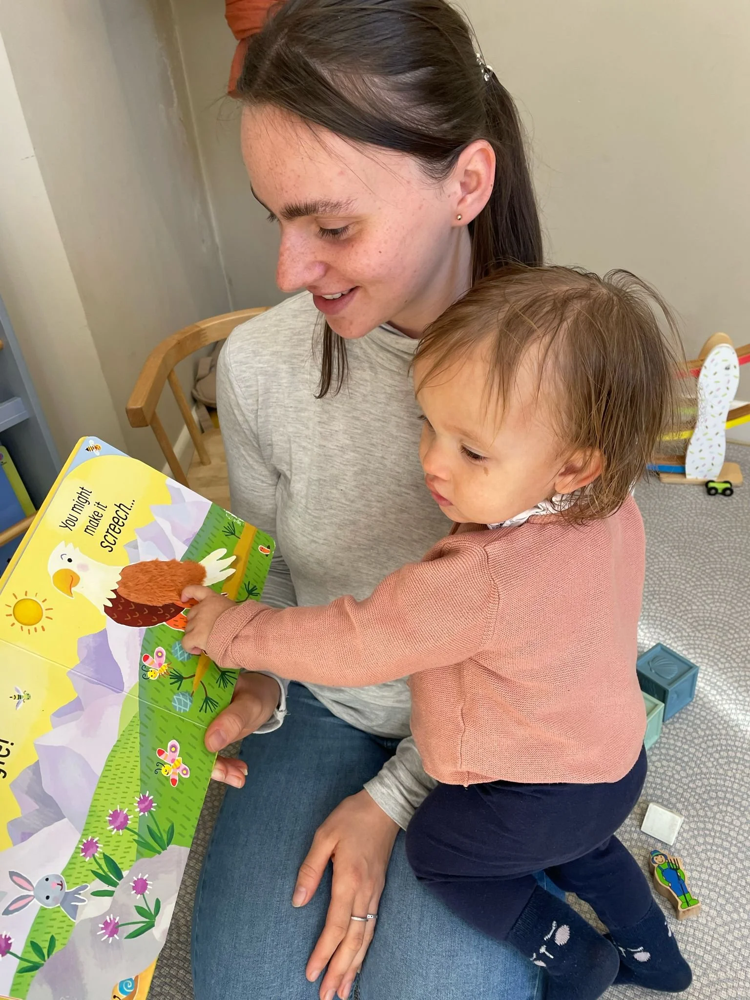 A woman with brown hair and a young girl with light brown hair sitting together, looking at a colorful children's book. The woman is smiling, and the girl is pointing at the book. They are indoors, seated on a blue cushion, with toys and a picture bo
