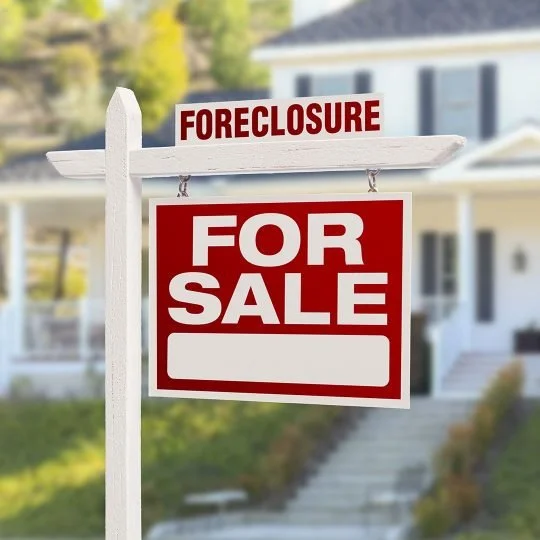 A house for sale sign with a 'Foreclosure' notice, in front of a residential home with greenery and a clear sky.