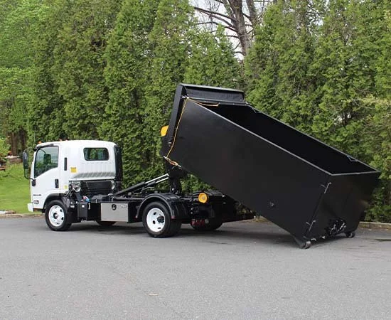 A white bucket truck with a black dump bed raised at an angle, parked on the side of a paved road with green trees in the background.