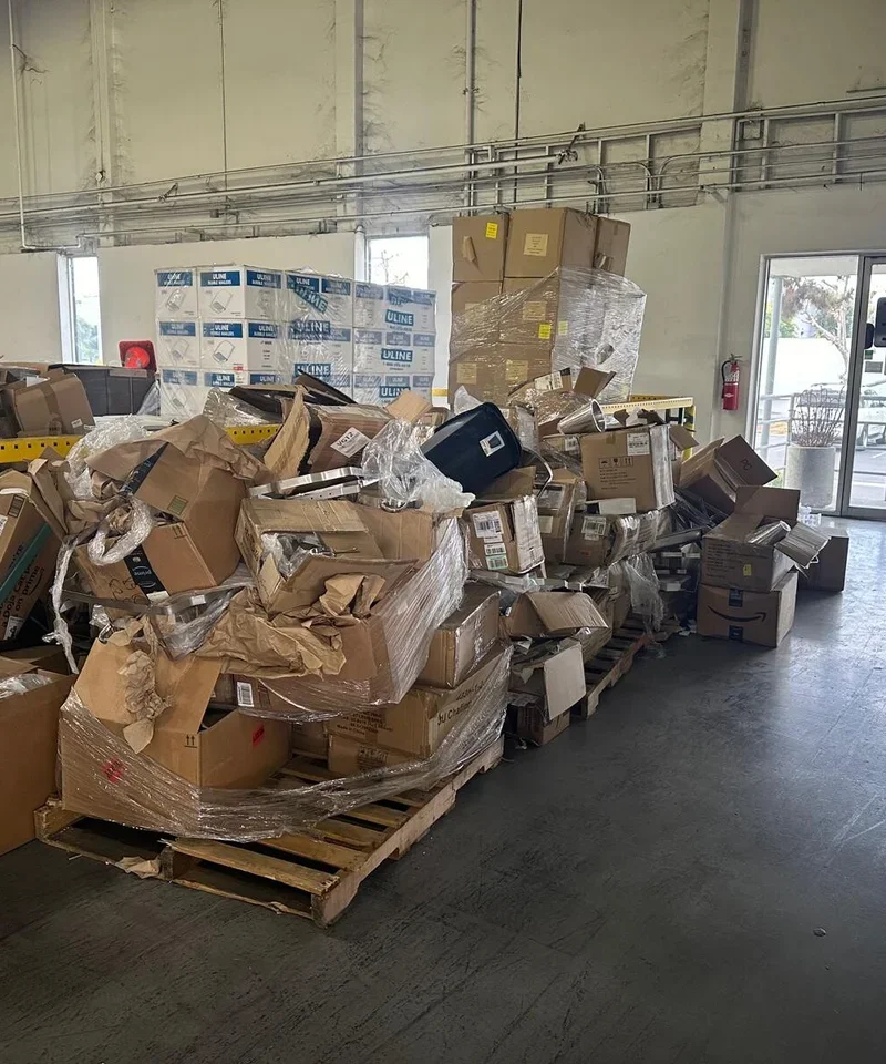A large pile of cardboard boxes and packaging materials on wooden pallets inside a warehouse or storage facility, with windows and a fire extinguisher visible in the background.