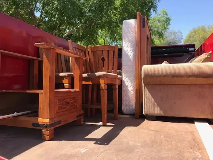 Outdoor scene with wooden chairs, a mattress, a wooden headboard, and a beige sofa on a deck with trees and a bright sky in the background.