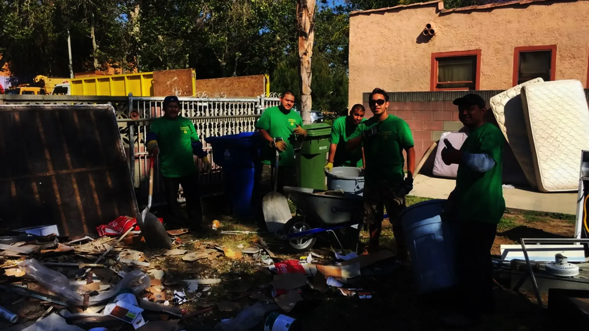 Group of five people in green shirts cleaning debris outside a building, with mattresses and household items, some garbage bins, and a fence in the background.