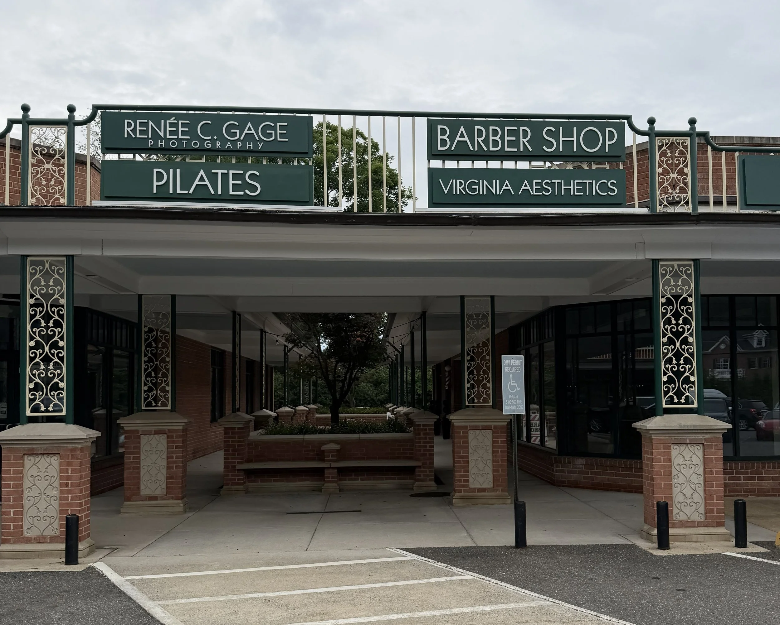 The image shows a building with multiple signs on the upper level. The signs include 'Renée C. Gage Photography', 'Pilates', 'Barber Shop', and 'Virginia Aesthetics'. The building features decorative metalwork on the columns, brick walls, and large windows. There is a small parking lot in front with marked spaces and a few black bollards. The sky above is cloudy with an overcast appearance.