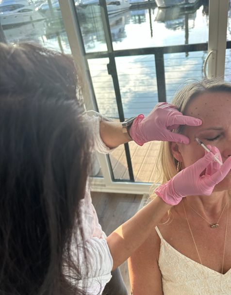 A woman receives a cosmetic injection from a medical professional wearing pink gloves at a clinic near a window overlooking a dock with boats.