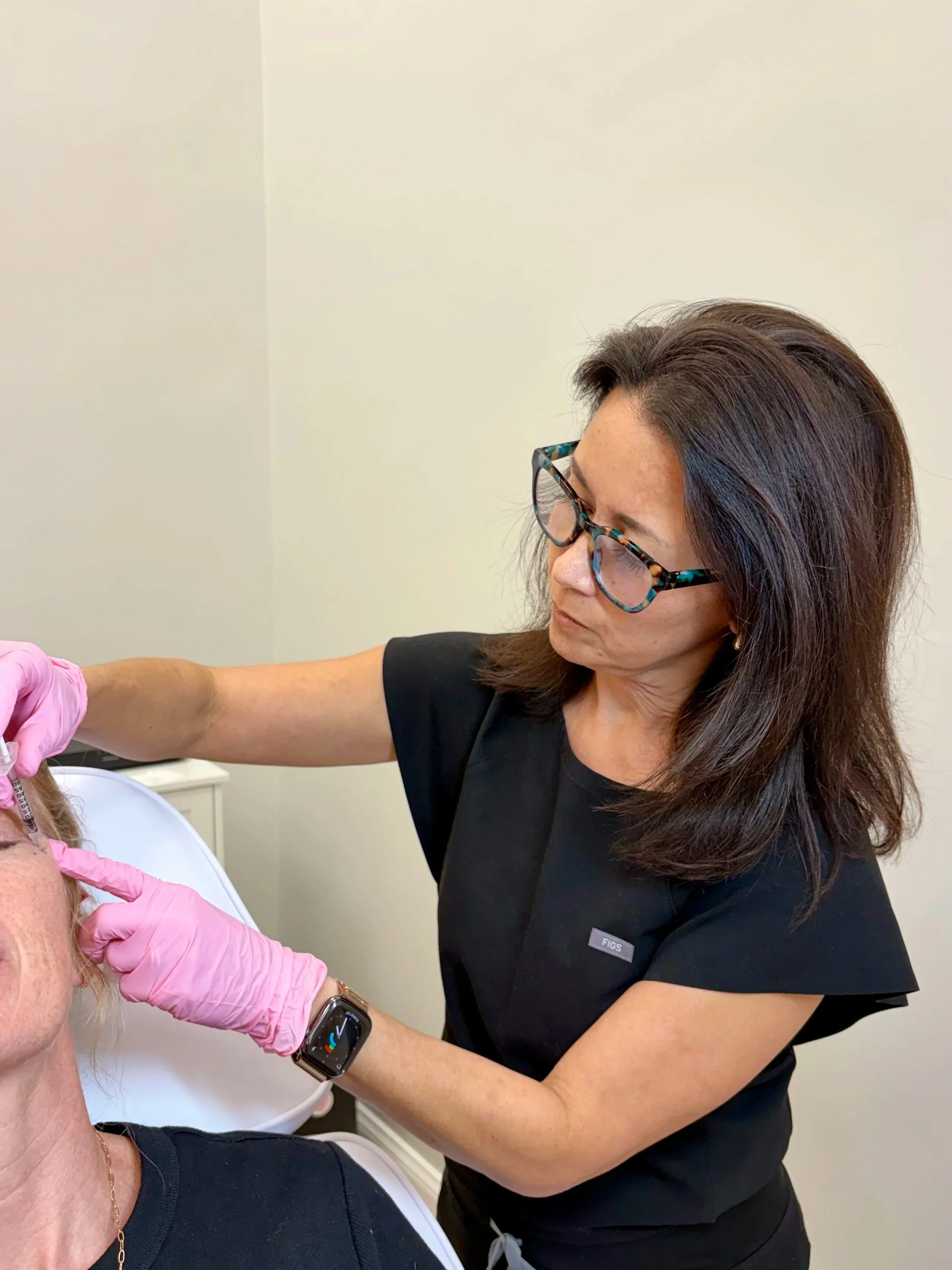 A woman with long brown hair and glasses is administering a vaccine to an older woman with blonde hair, wearing a black shirt with pink gloves in a clinical setting.