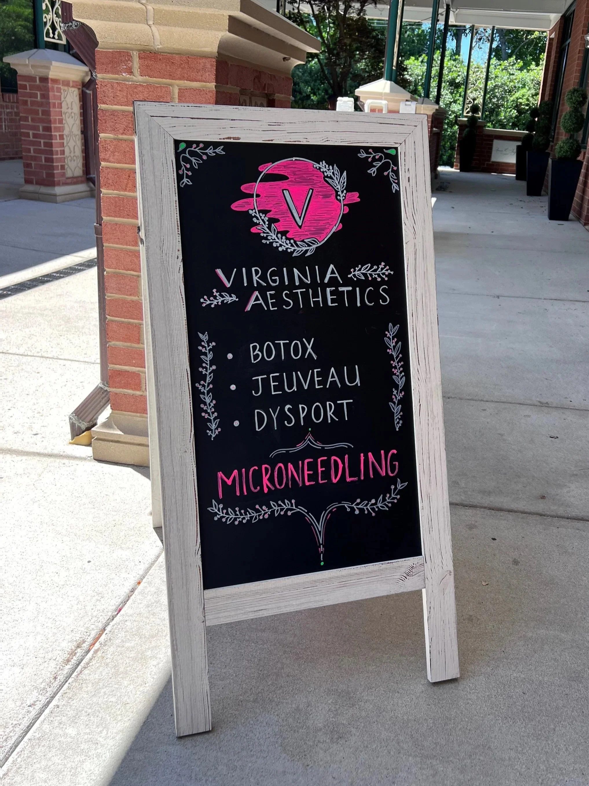 A chalkboard sign outside a business named Virginia Aesthetics. The sign lists services including botox, jeuveau, dysport, and microneedling, decorated with floral and botanical designs.