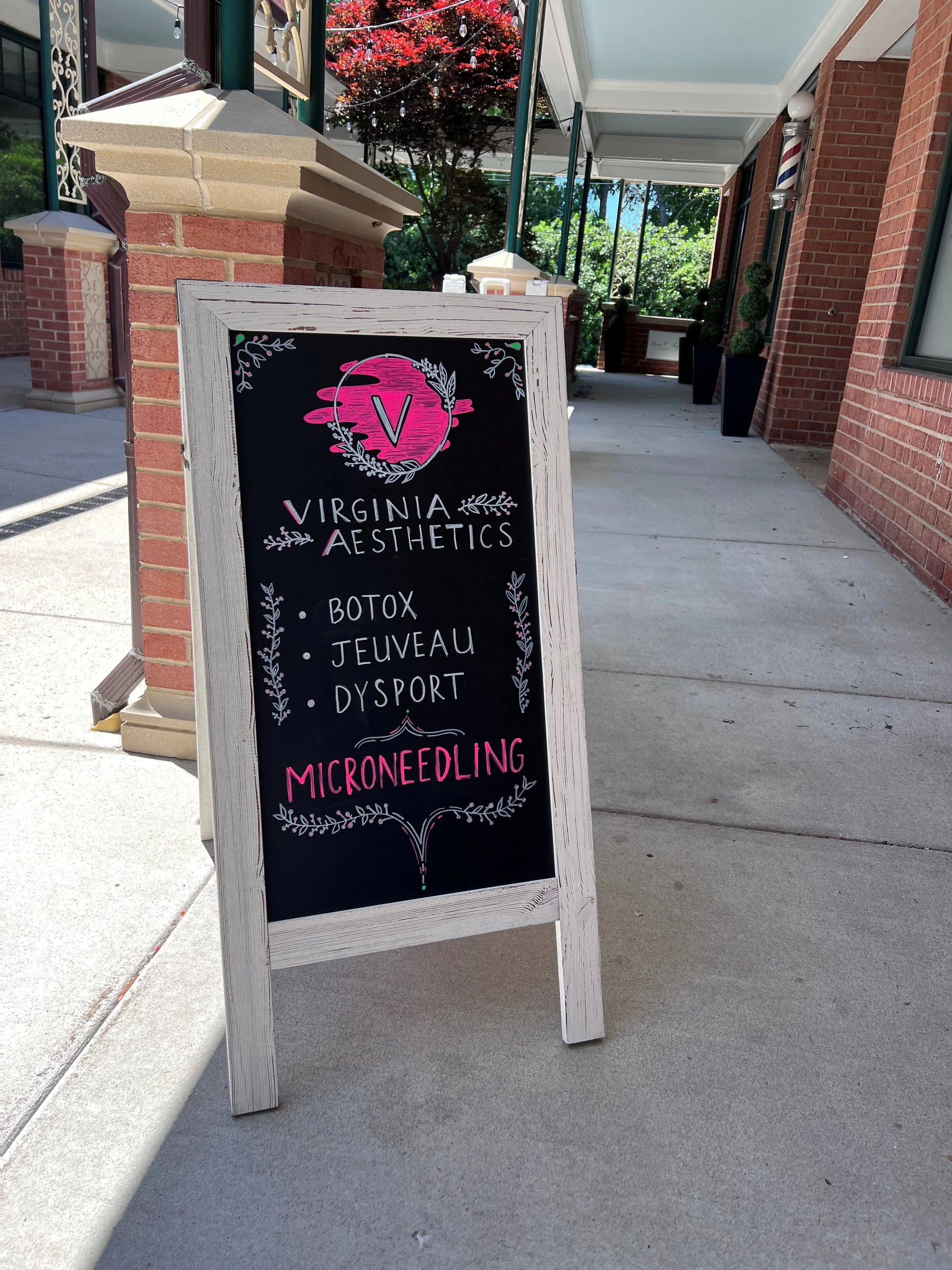 Outdoor sidewalk chalkboard sign for Virginia Aesthetics listing services such as Botox, Jeuveau, Dysport, and Microneedling, with trees and brick building in the background.