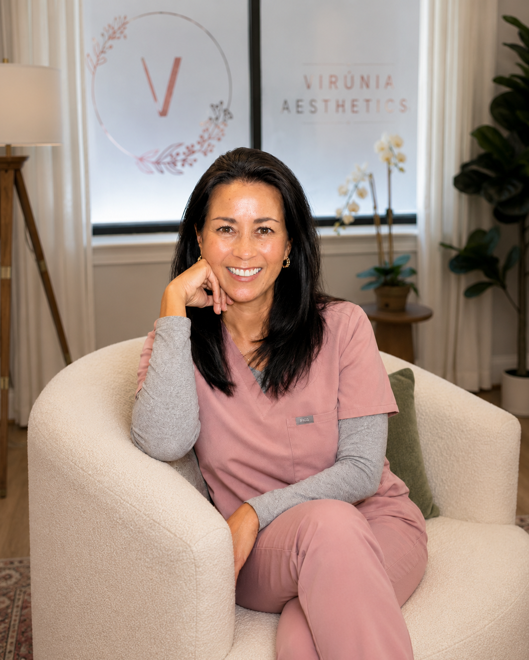 A woman sitting on a beige chair in a medical or aesthetic clinic, smiling at the camera. She is wearing pink scrubs with gray sleeves and has dark hair. Behind her, there are two windows, a sign with a logo that says 'Virūnia Aesthetics,' and a potted orchid plant.