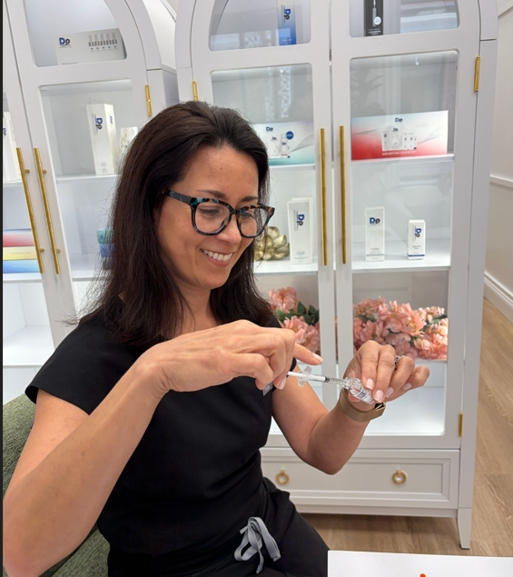 A woman with dark hair and glasses preparing a syringe indoors, with a display cabinet containing skincare or cosmetic products and pink flowers in the background.