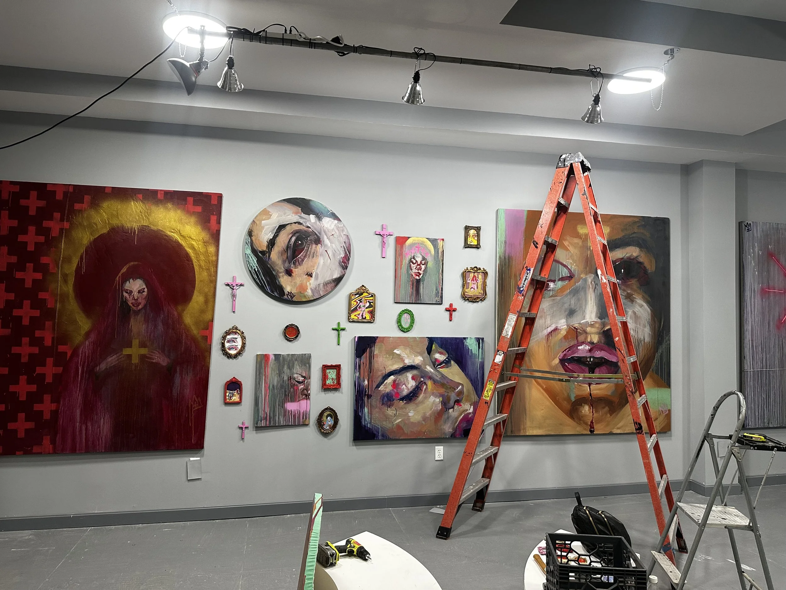 An art gallery wall featuring several colorful abstract paintings and framed images, with a red step ladder set up in front. There are tools, a black basket, and a small step stool on the gray tiled floor, indicating ongoing setup or renovation.