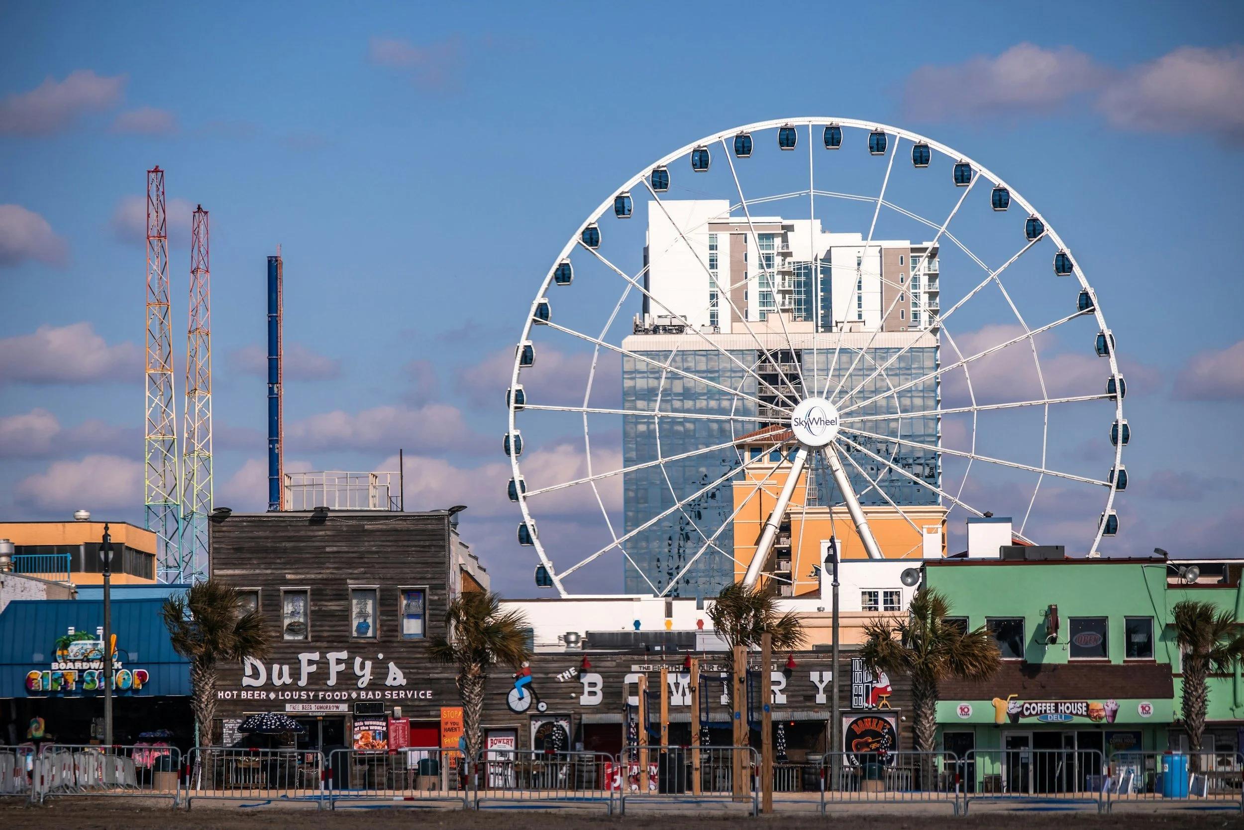 A cityscape featuring a large Ferris wheel in the foreground, with high-rise buildings and colorful shops beneath, under a blue sky with scattered clouds.