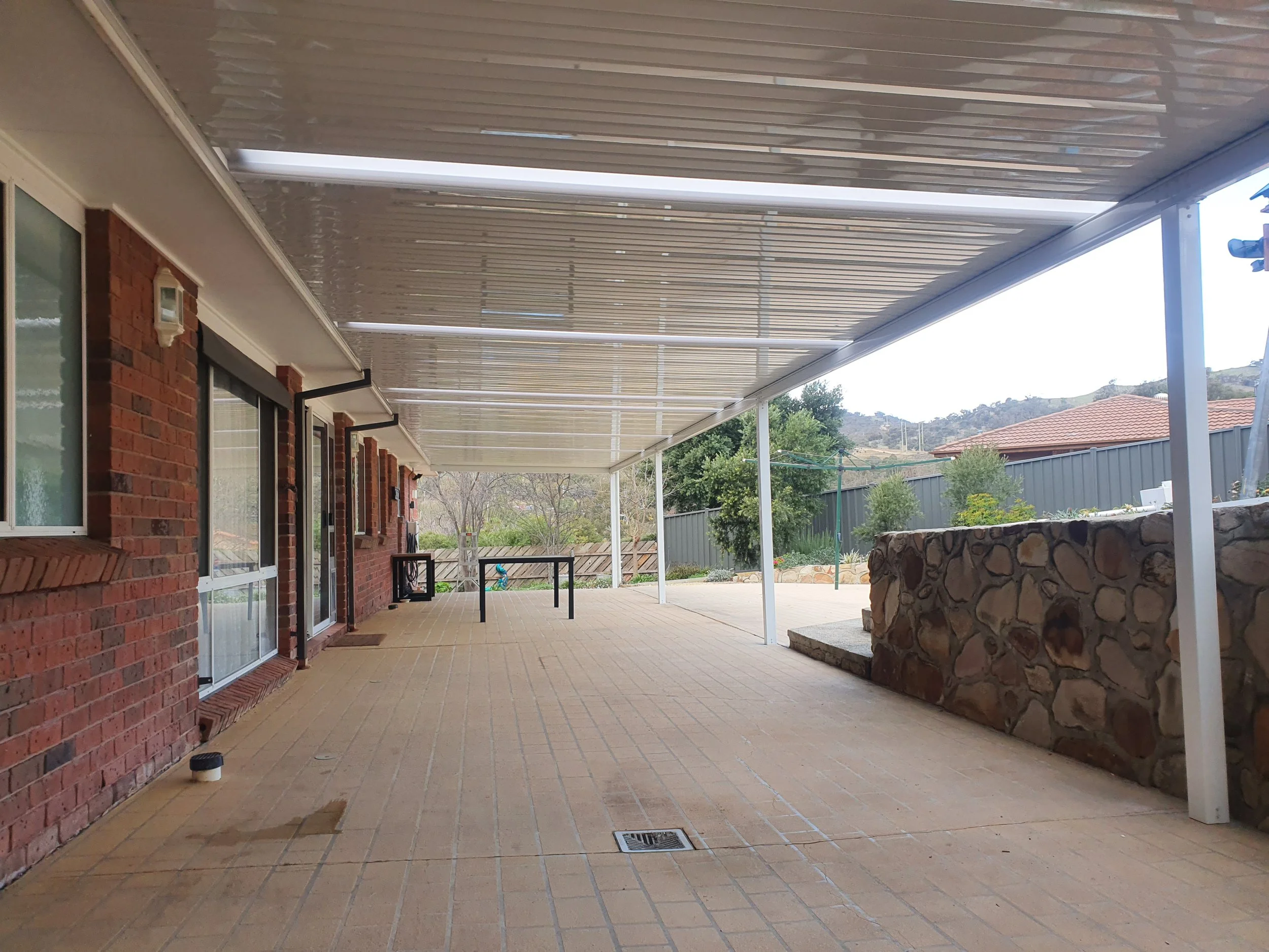 Covered patio area attached to a brick house, with a tiled floor and a raised stone feature. Overhead, there is a translucent roof with white support beams, and a landscape with trees and a sloped hill in the background.