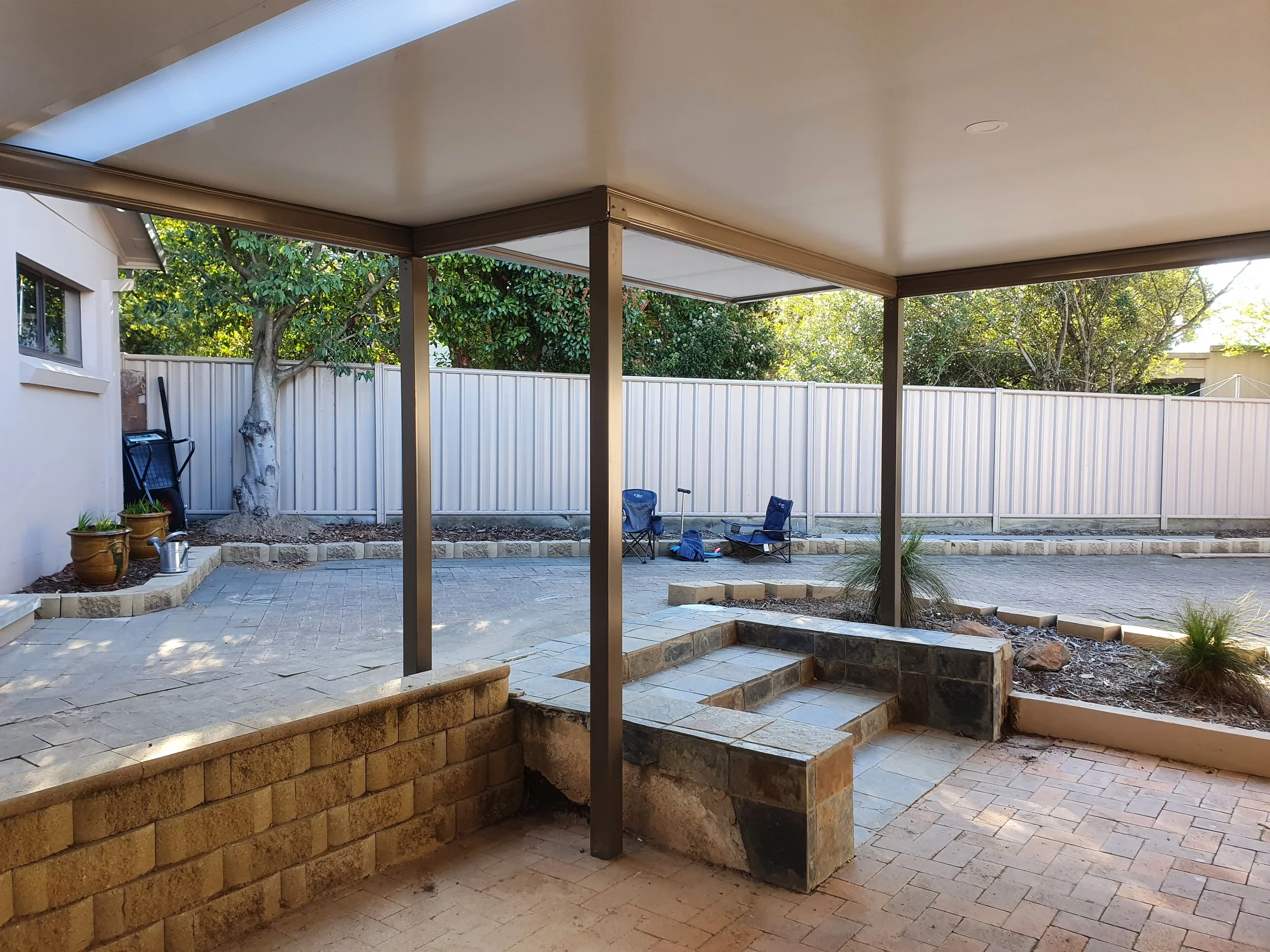 A backyard patio area featuring a stone staircase, potted plants, a white fence, a large tree, and outdoor chairs. The area is partially shaded by a covered patio.
