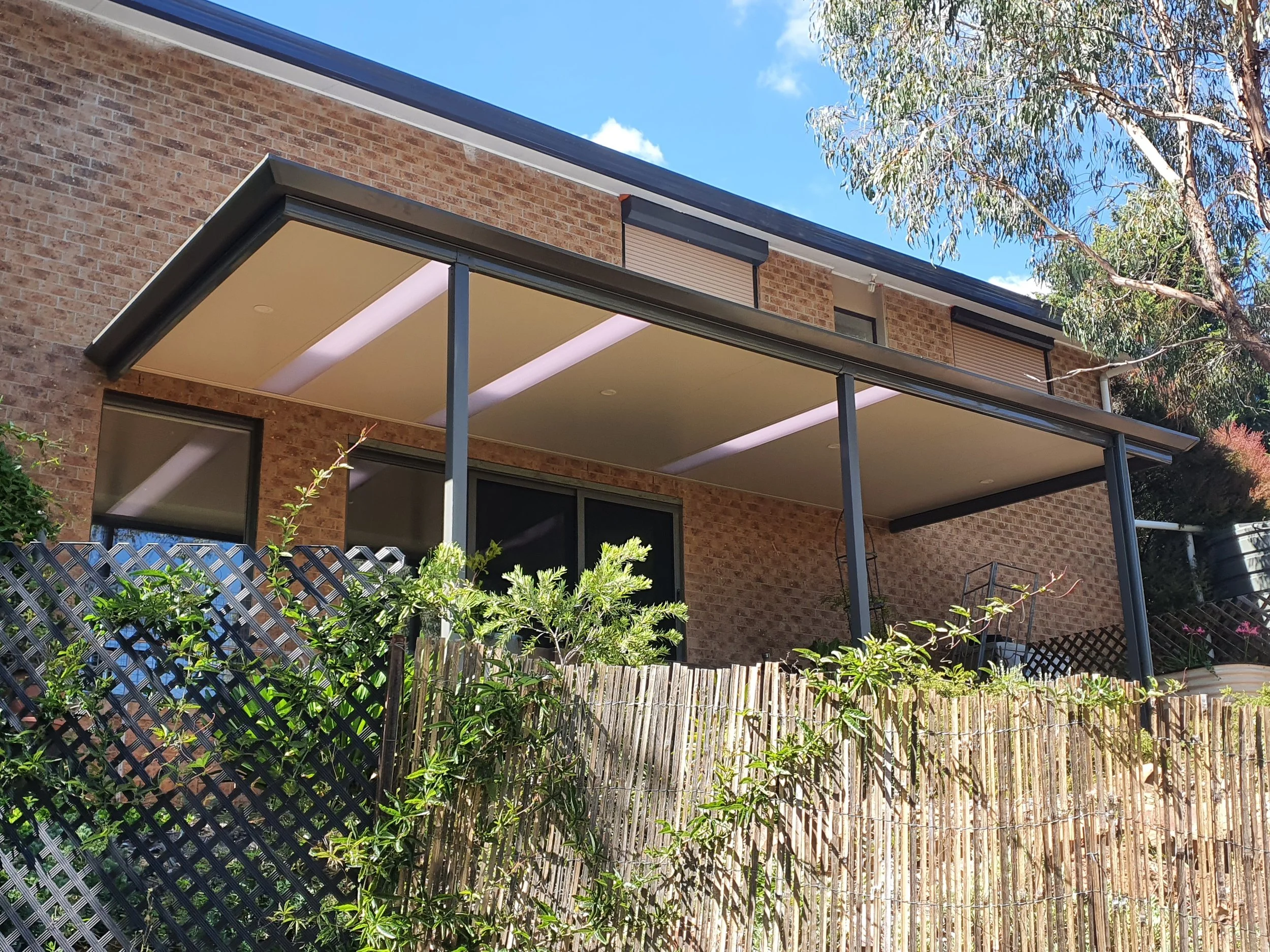 A view of a brick house with a patio covered by a beige ceiling and surrounded by a wooden fence and green plants, with a large tree and blue sky in the background.