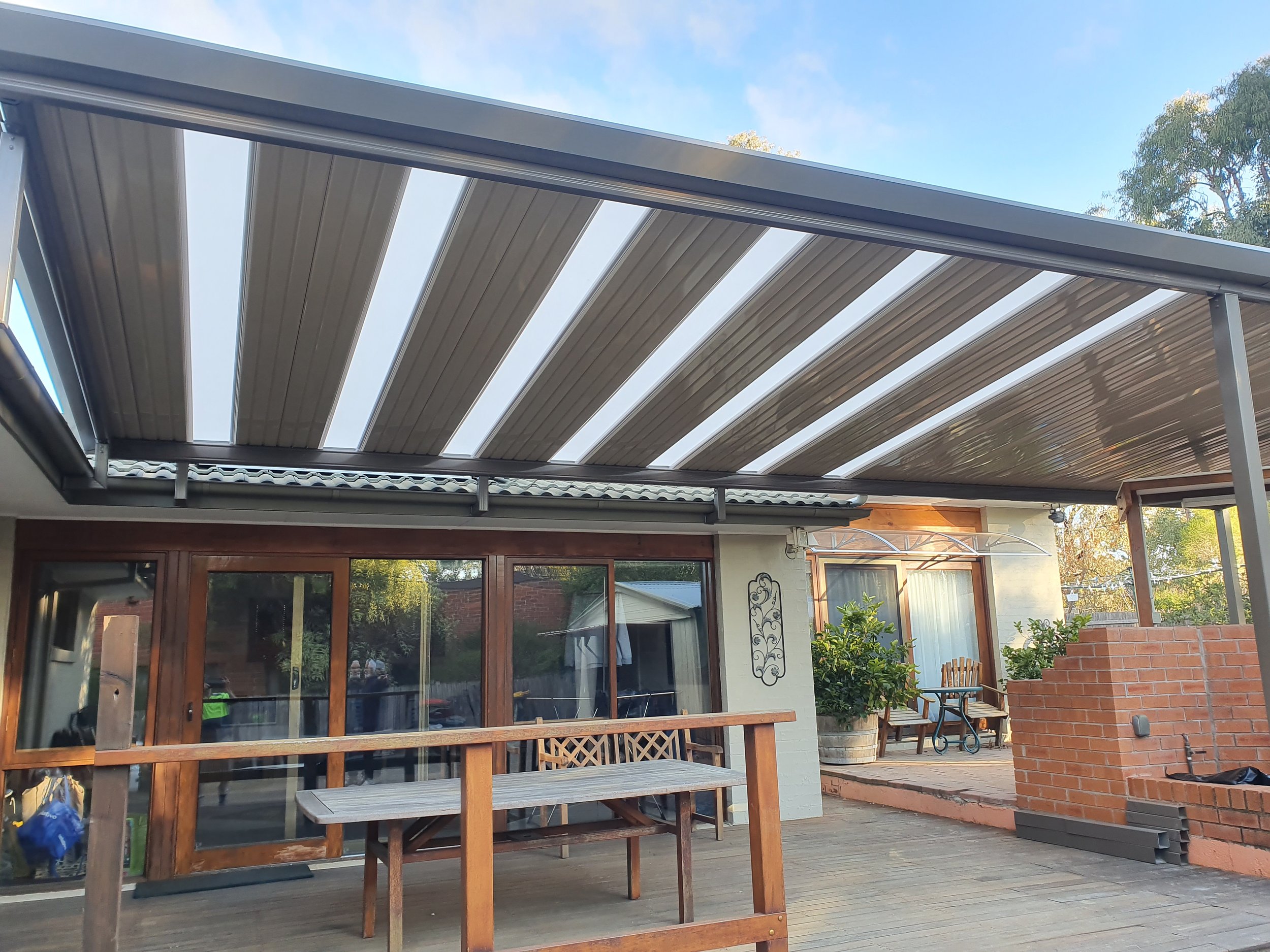 Backyard patio area featuring a covered pergola with metal framing, wooden railing, sliding glass doors, and outdoor furniture, including a wooden table, chairs, and potted plants, with trees and a blue sky in the background.