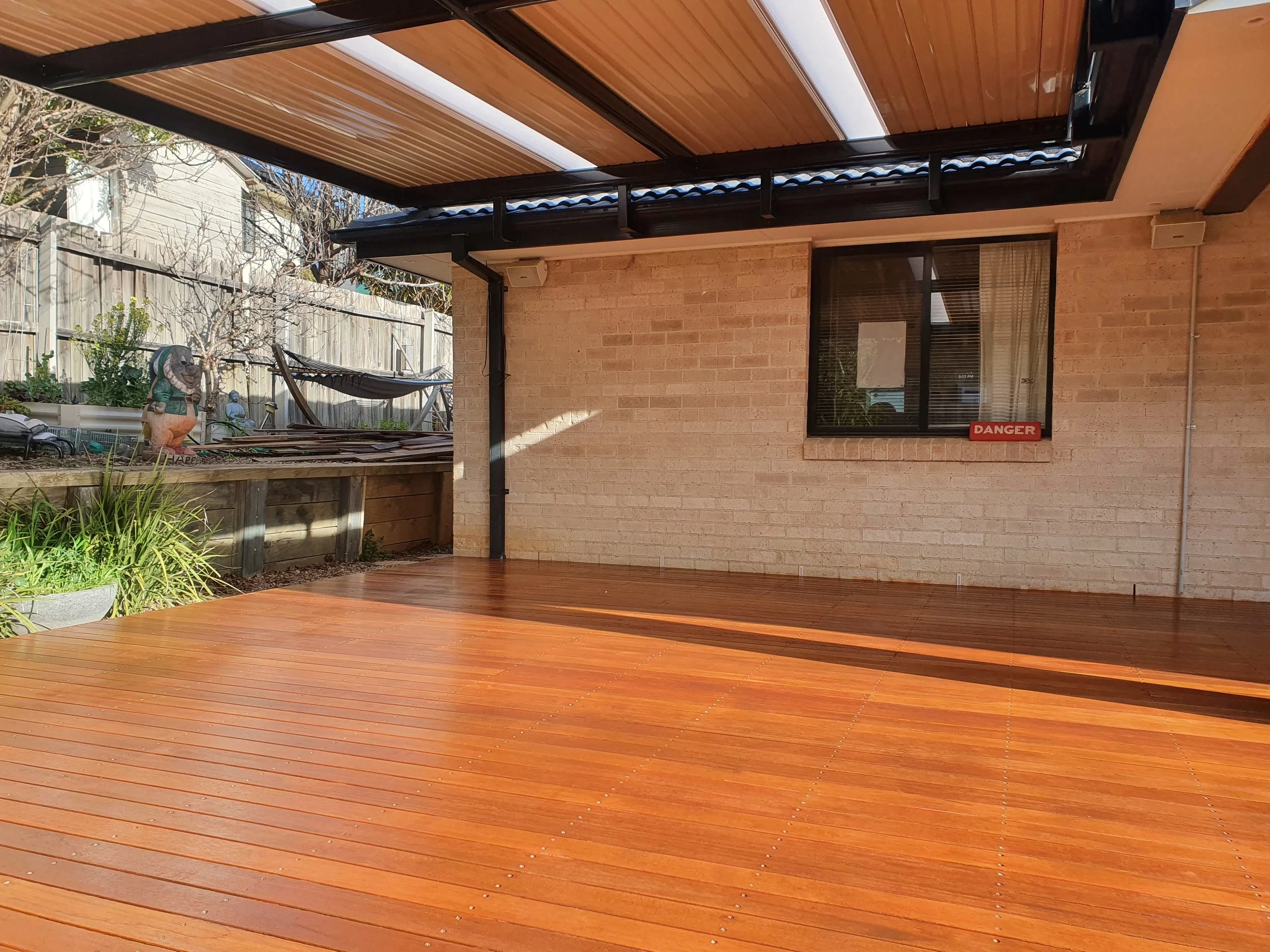 Backyard patio area with a wooden deck, a house wall with a window, garden plants, a garden statue, a hammock, and a modern roof overhang.