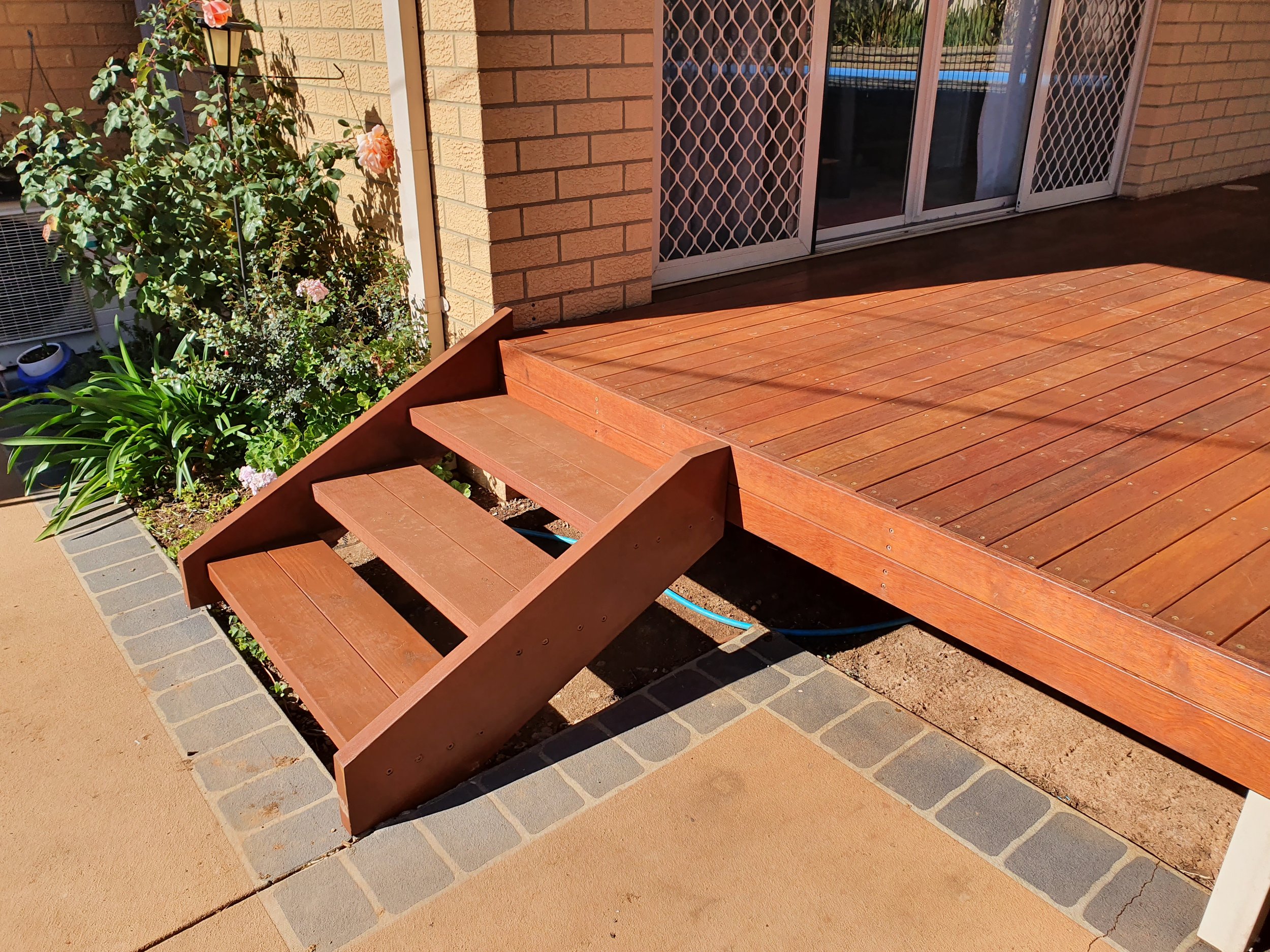 A wooden deck with steps next to a brick wall and a sliding glass door. To the left, there are flowering plants with pink roses and green leaves, partially shaded by sunlight.